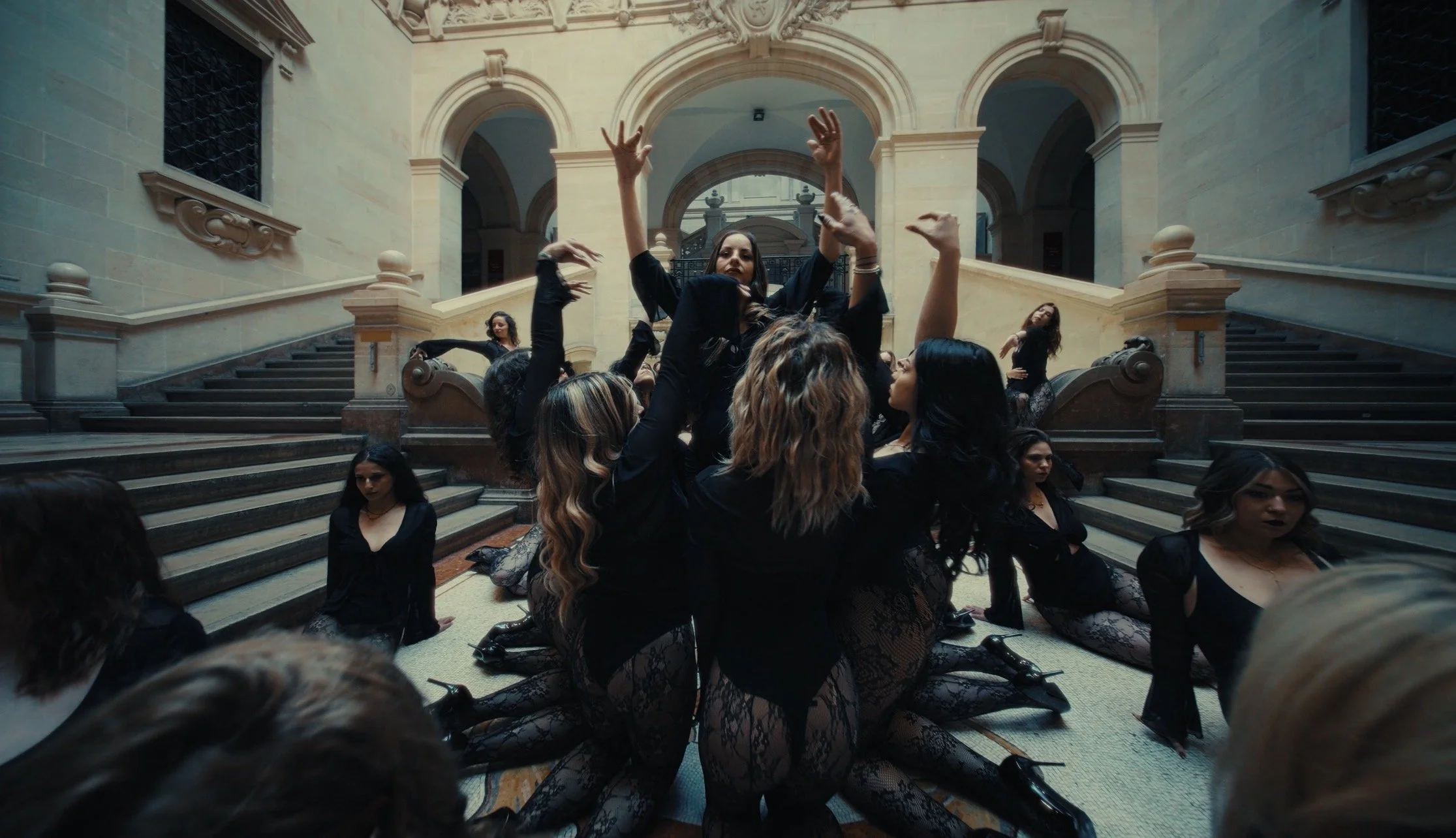 Group of women in black clothing and lace tights performing a dance or yoga pose on staircase in a grand, historic building with arches and decorative architecture.