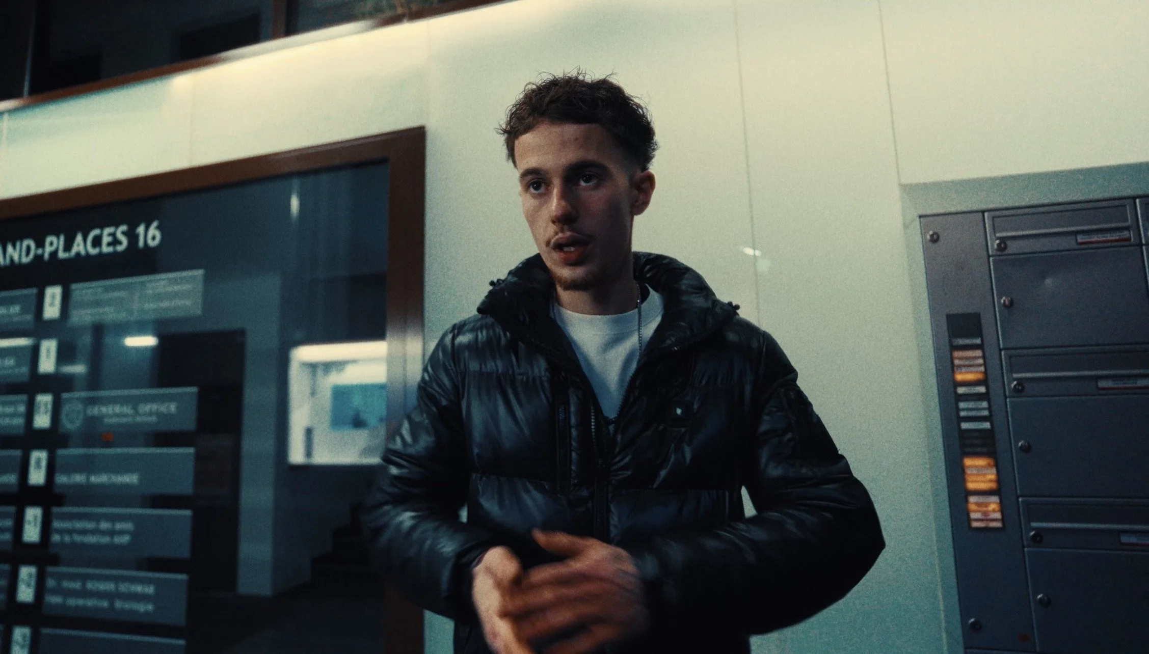 A young man in a black shiny jacket standing at an airport check-in area with lockers behind him.