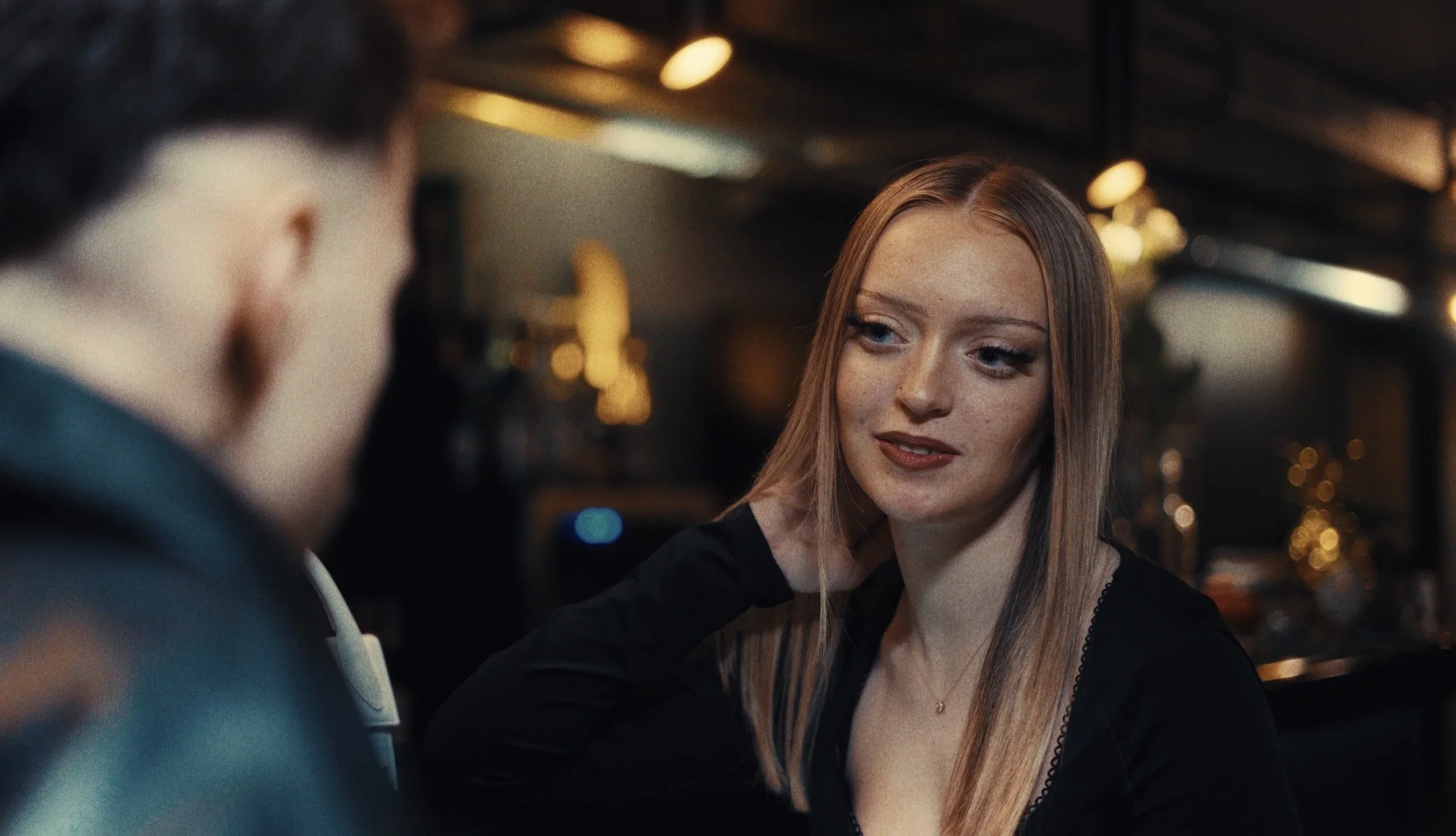 A woman with red hair and a black top looking at a man with dark hair, who is partially blurred in the foreground, inside a dimly lit restaurant or bar with warm lighting and blurred background decor.