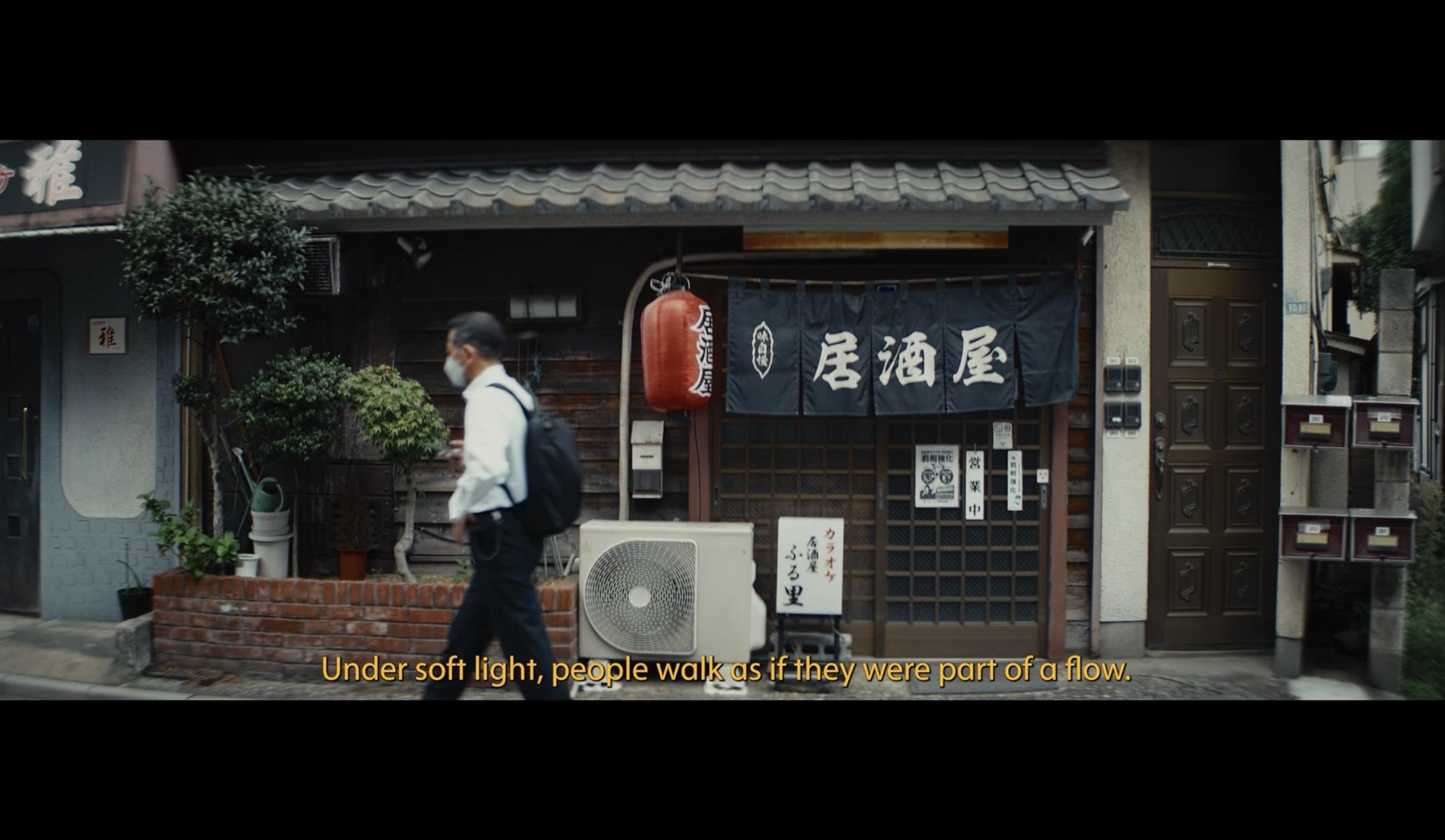 A man in a business suit and face mask walking past a traditional Japanese restaurant with a red paper lantern and dark blue curtain with white Japanese characters. Subtitles read, 'Under soft light, people walk as if they were part of a flow.'