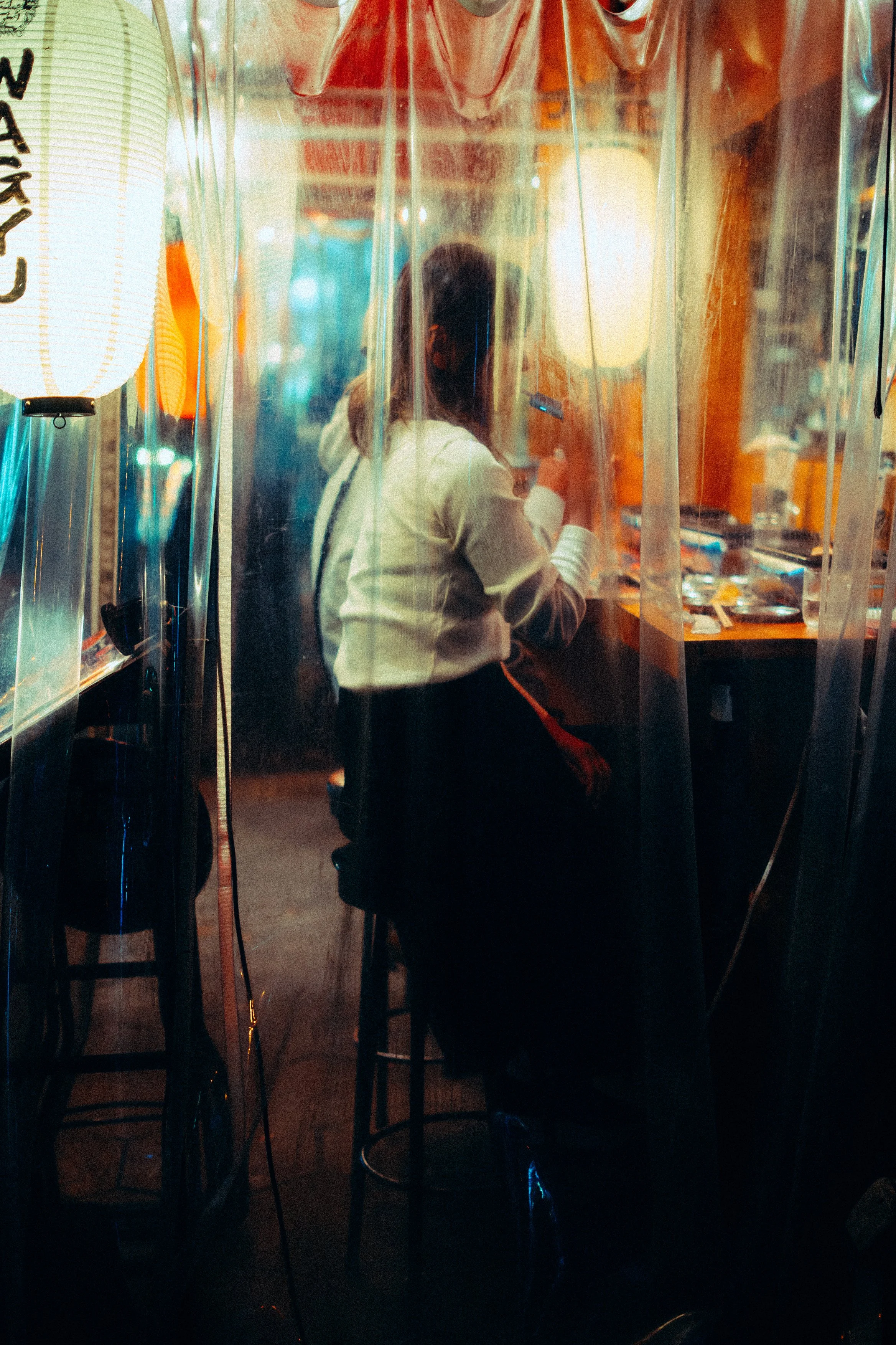 A woman sitting between a transparent plastic partition inside a restaurant or cafe at night, looking at her phone, with dim warm lights and Asian-inspired decor.