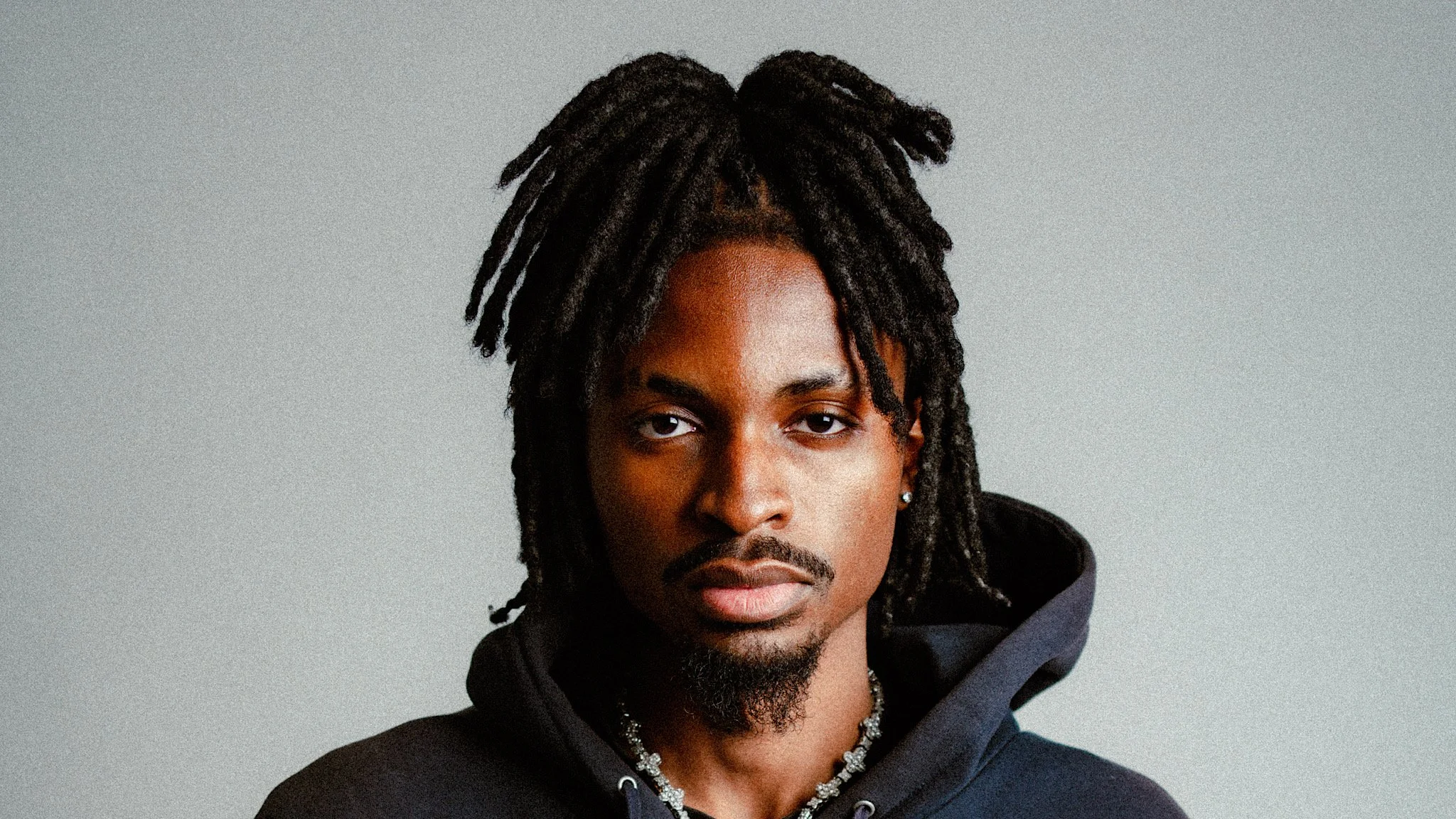 Close-up of a young man with medium skin tone, dreadlocks, goatee, wearing a black hoodie and jewelry, looking directly at the camera against a plain background.