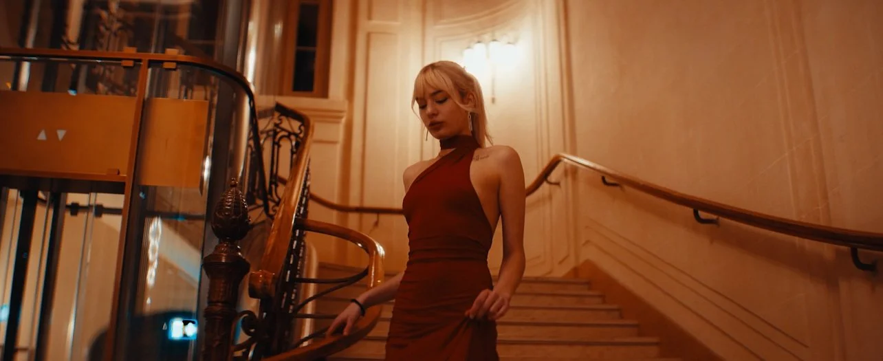 A young woman with blonde hair in a high-neck dress walking down a wooden staircase in an elegant, warmly lit interior.