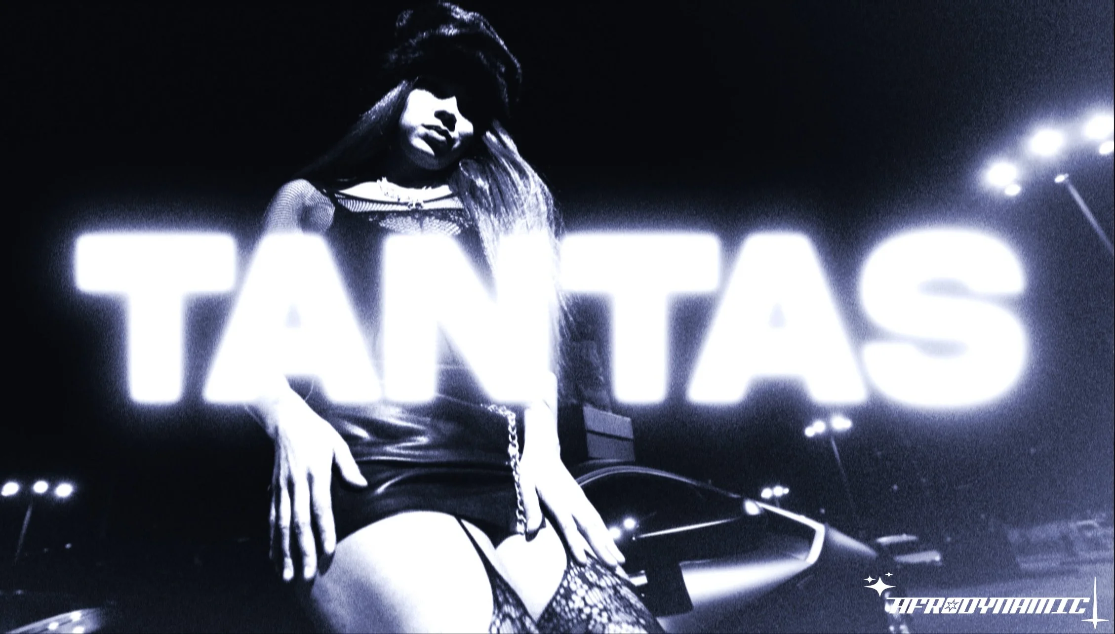 A woman wearing fishnet stockings, a leather skirt, and a top with mesh details, standing in front of a neon sign that says 'TANTAS' in an urban setting at night.