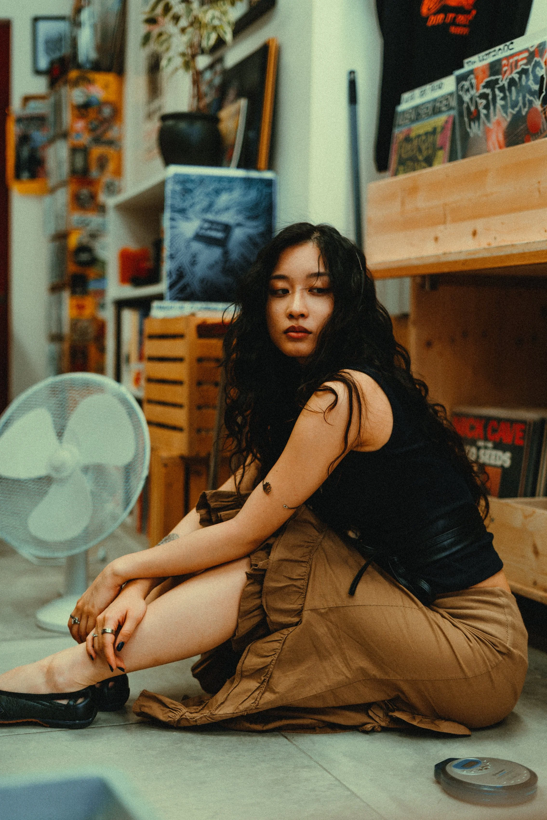 A woman with black curly hair sitting on the floor in a room with art and posters on the walls. She is dressed in a black shirt and tan cargo pants, resting her arms on her knees, looking to the side. A small fan is visible in the foreground, and the