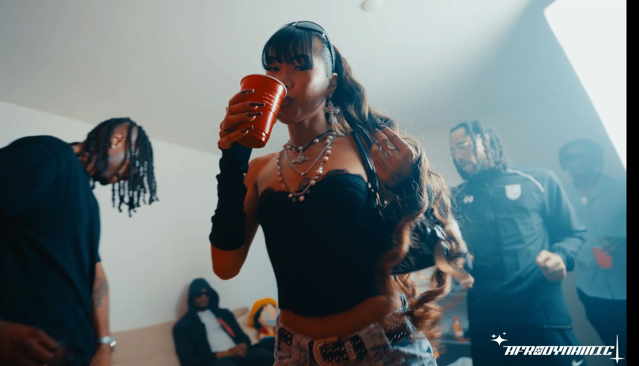 A woman with long wavy hair wearing jewelry, a black top, and patterned pants holds a red cup while drinking at a social gathering, with other people in the background.