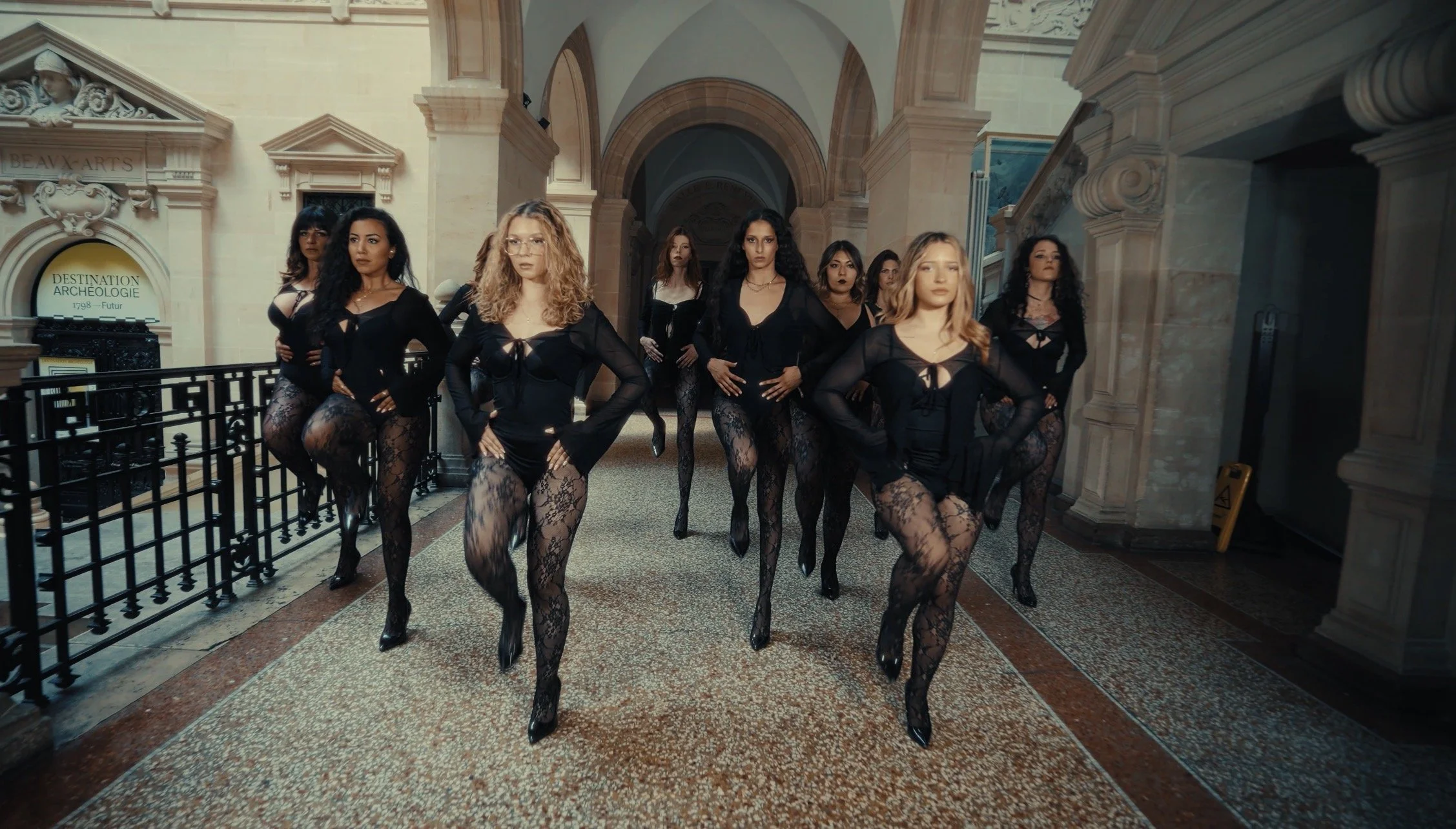 Group of nine women posing in elegant black lace outfits and high heels inside a historic building with arched ceilings and ornate architectural details.