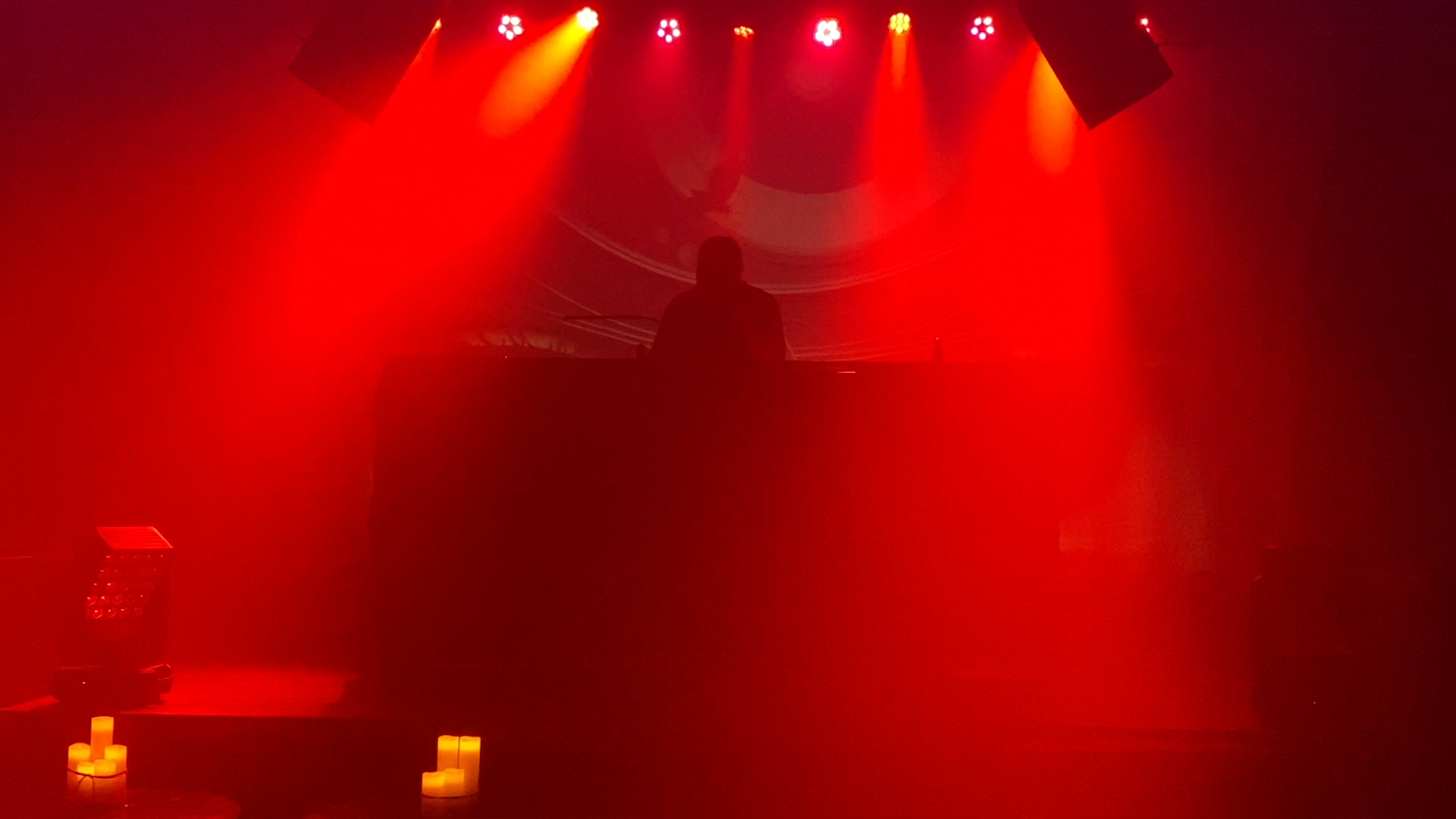 A DJ performing on stage with red and yellow lighting, silhouette of the DJ behind the equipment, and candles on the floor in front of the stage.