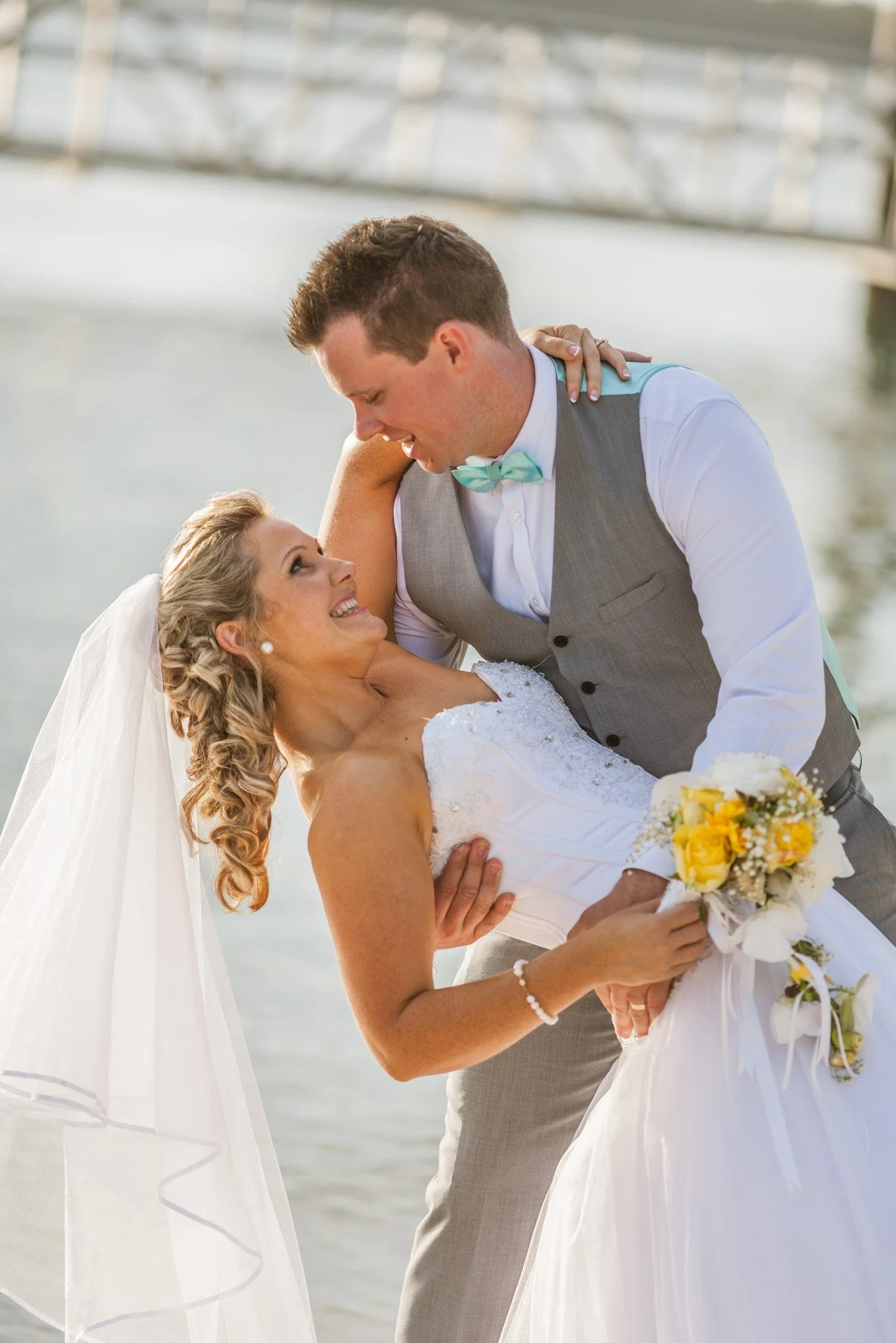 A bride and groom dancing outdoors near a body of water, with the groom dipping the bride and she holding a bouquet of yellow and white flowers.