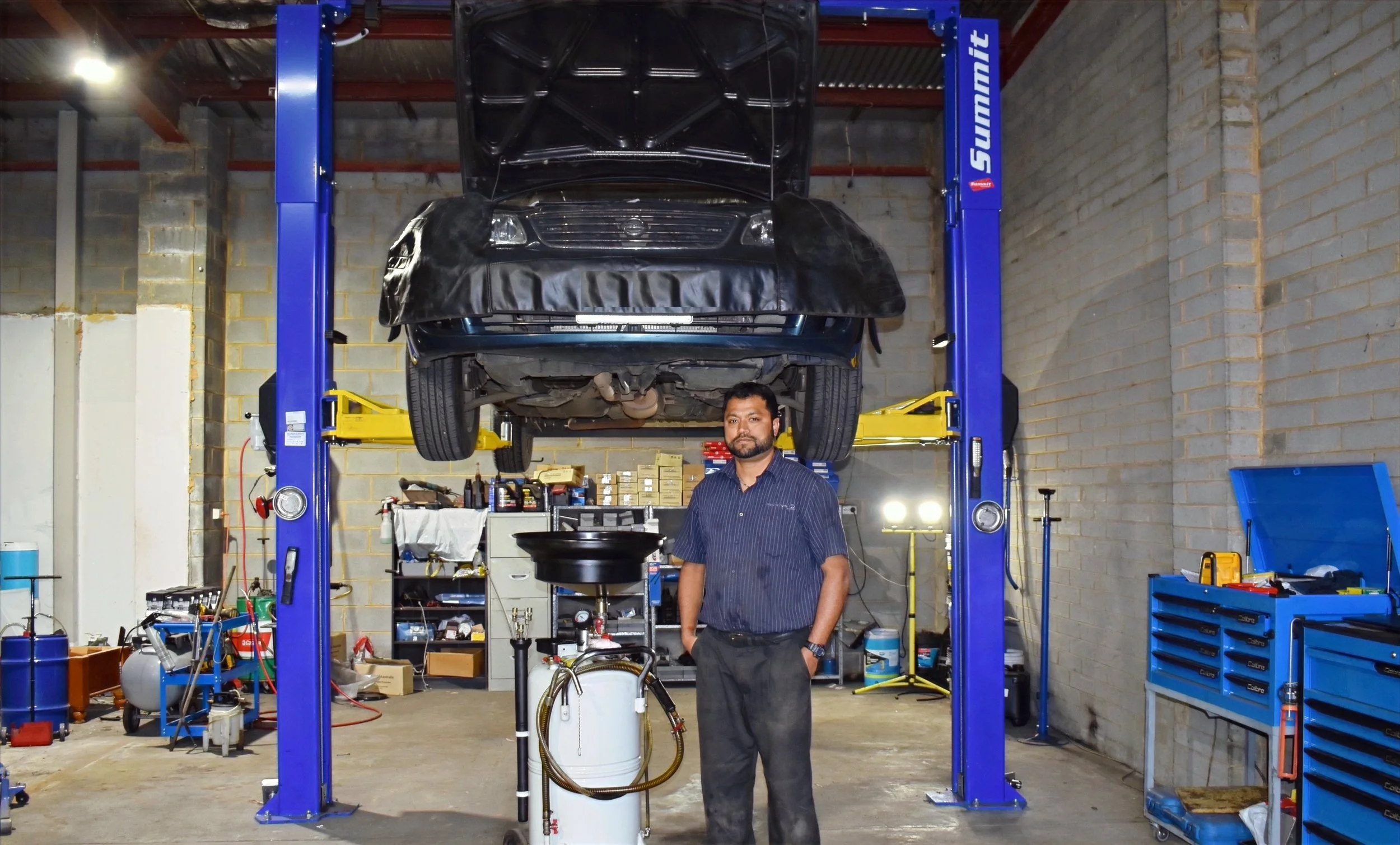 A man standing in an auto repair shop beneath a car lifted on a hydraulic lift. The car's hood is open, and there are various tools and equipment around.
