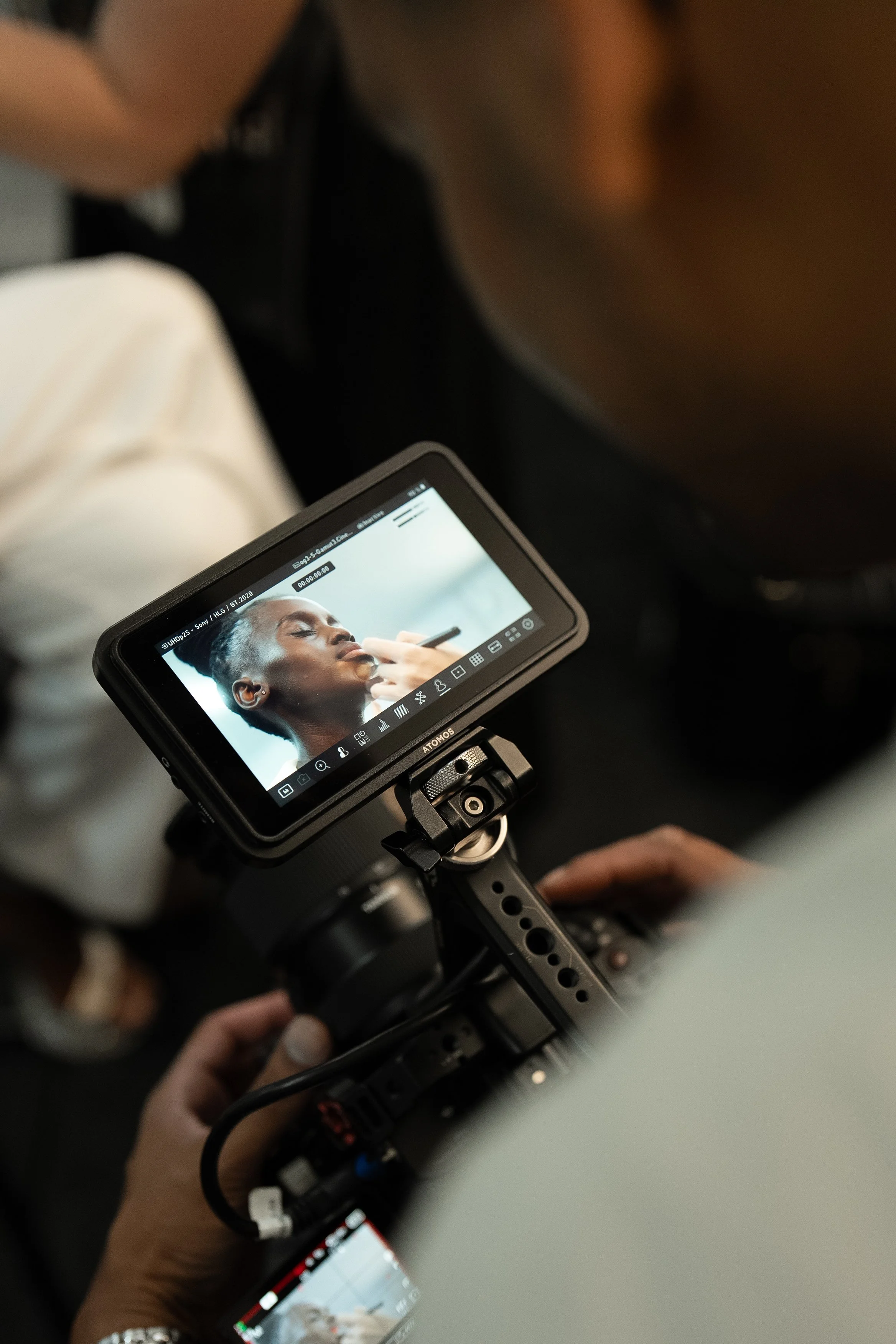 A camera monitor displays a woman with short hair applying makeup, seen from above during a photoshoot or filming.