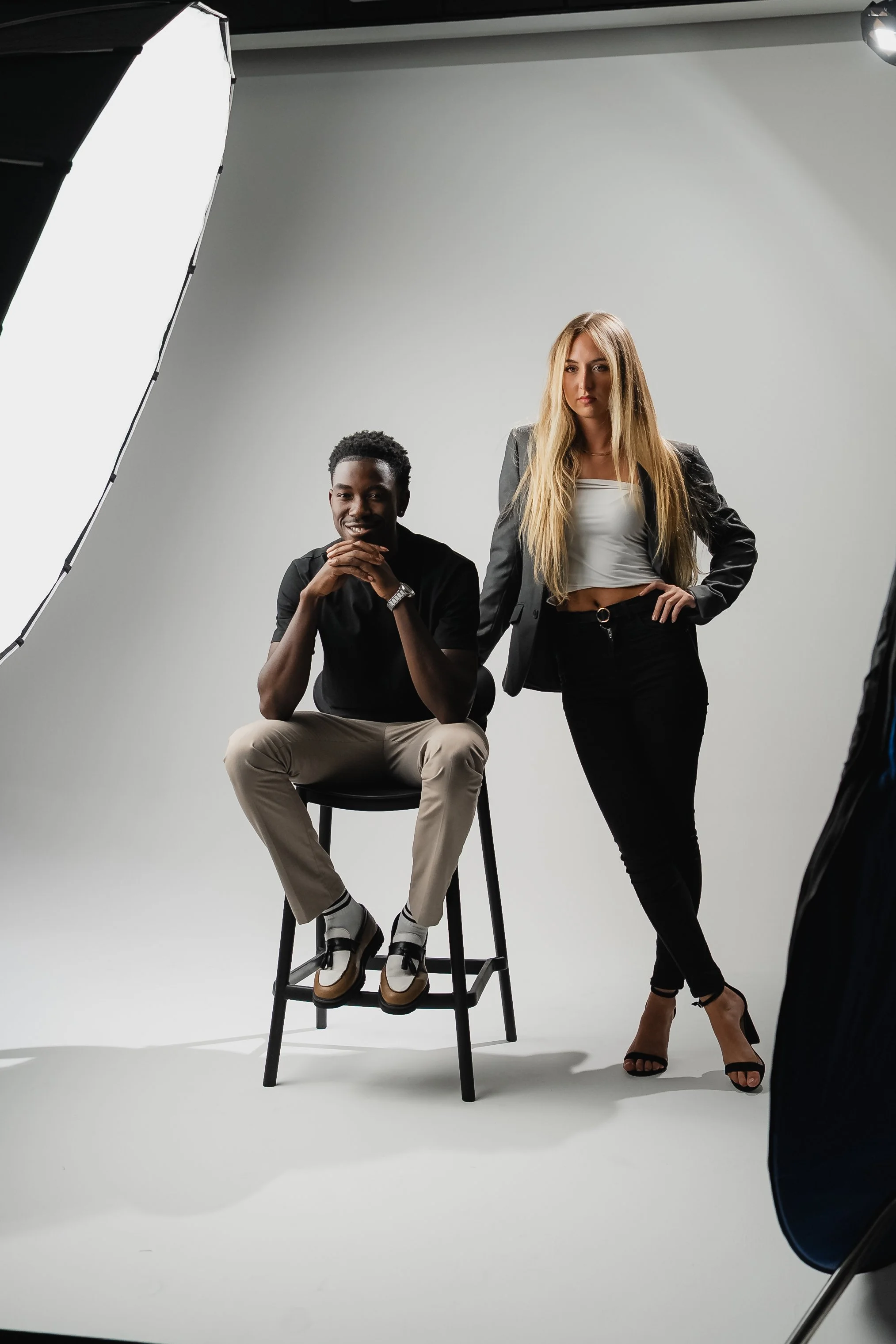 Two young women in a photography studio with a white backdrop. One is seated on a stool, smiling with hands clasped, wearing a black shirt and beige pants. The other stands beside her, hands on hips, with long blonde hair, wearing a black blazer, white crop top, and black pants.