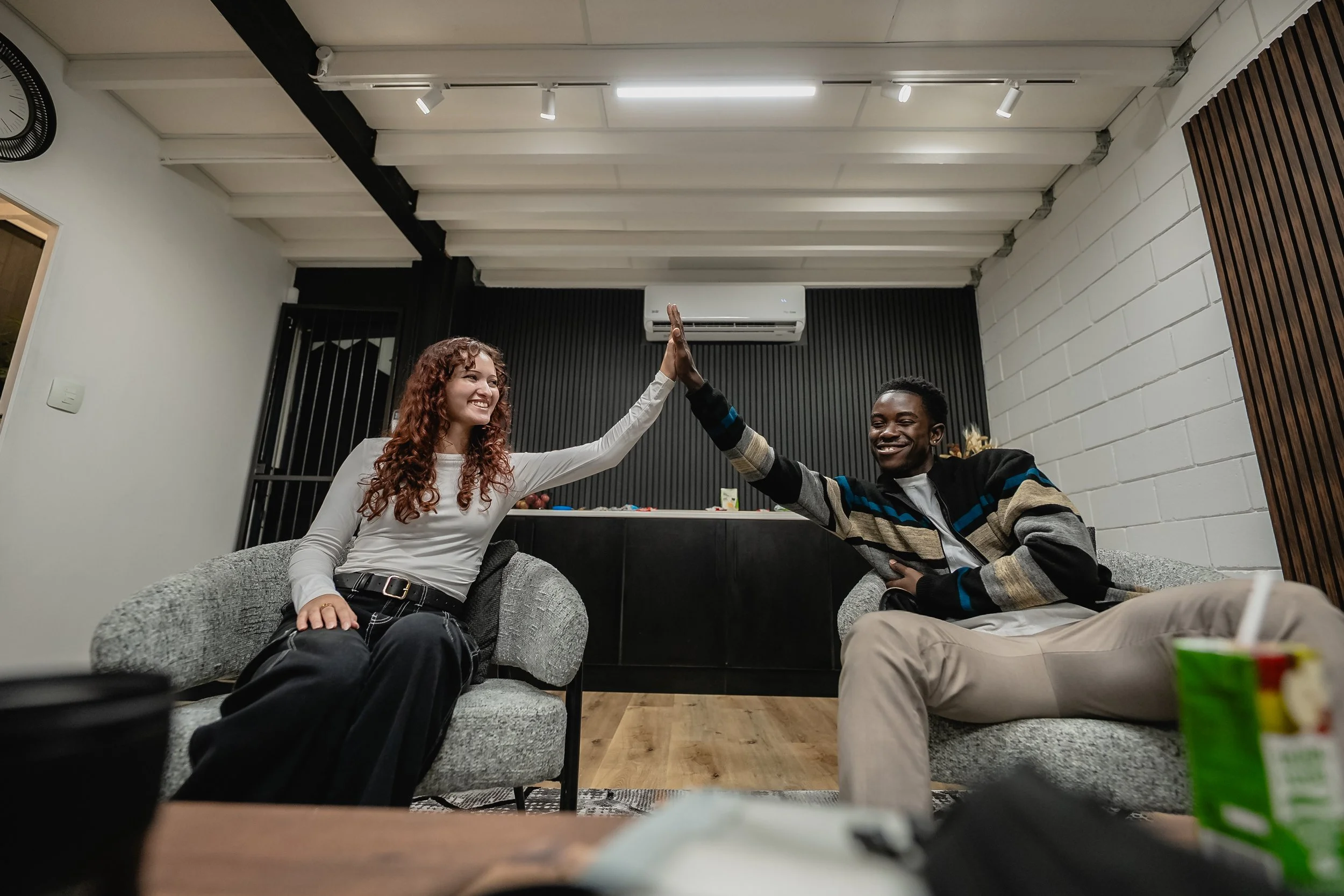 Two friends, a woman with curly red hair and a man with short black hair, sitting on sofas in a modern room, giving each other a high five and smiling.