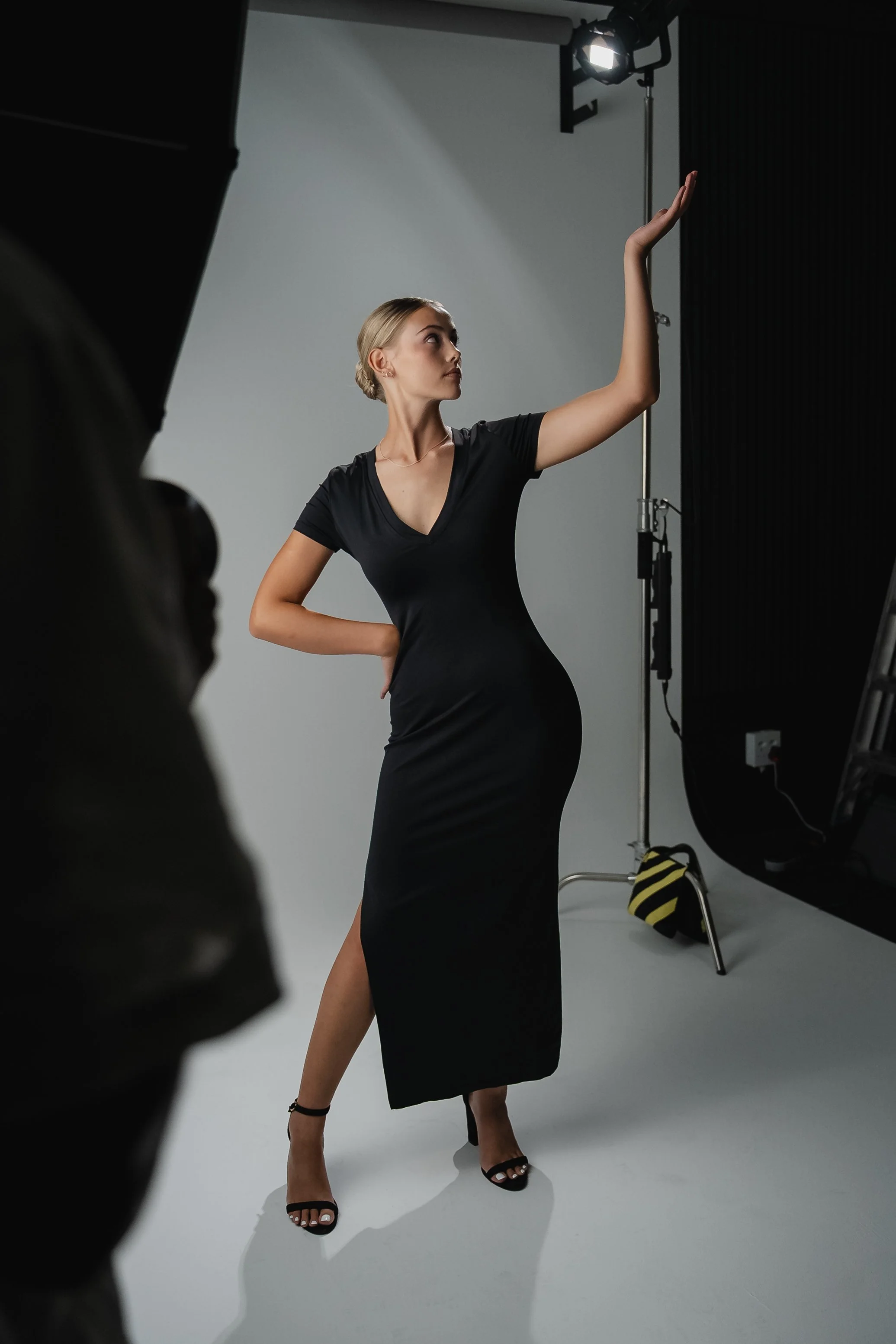 A woman in a black dress and high heels posing in a photography studio with a white backdrop and studio lights.