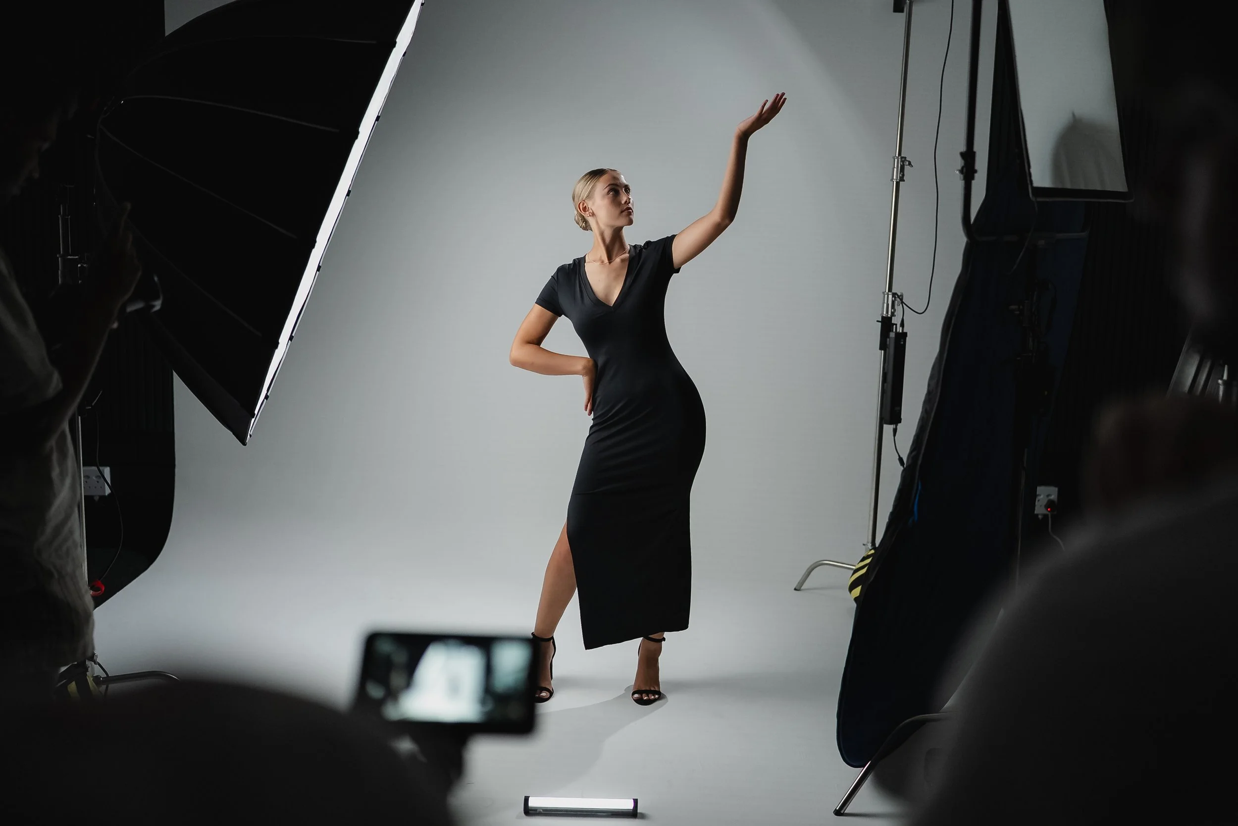 A woman posing in a black dress for a photo shoot in a photography studio with professional lighting and background equipment.
