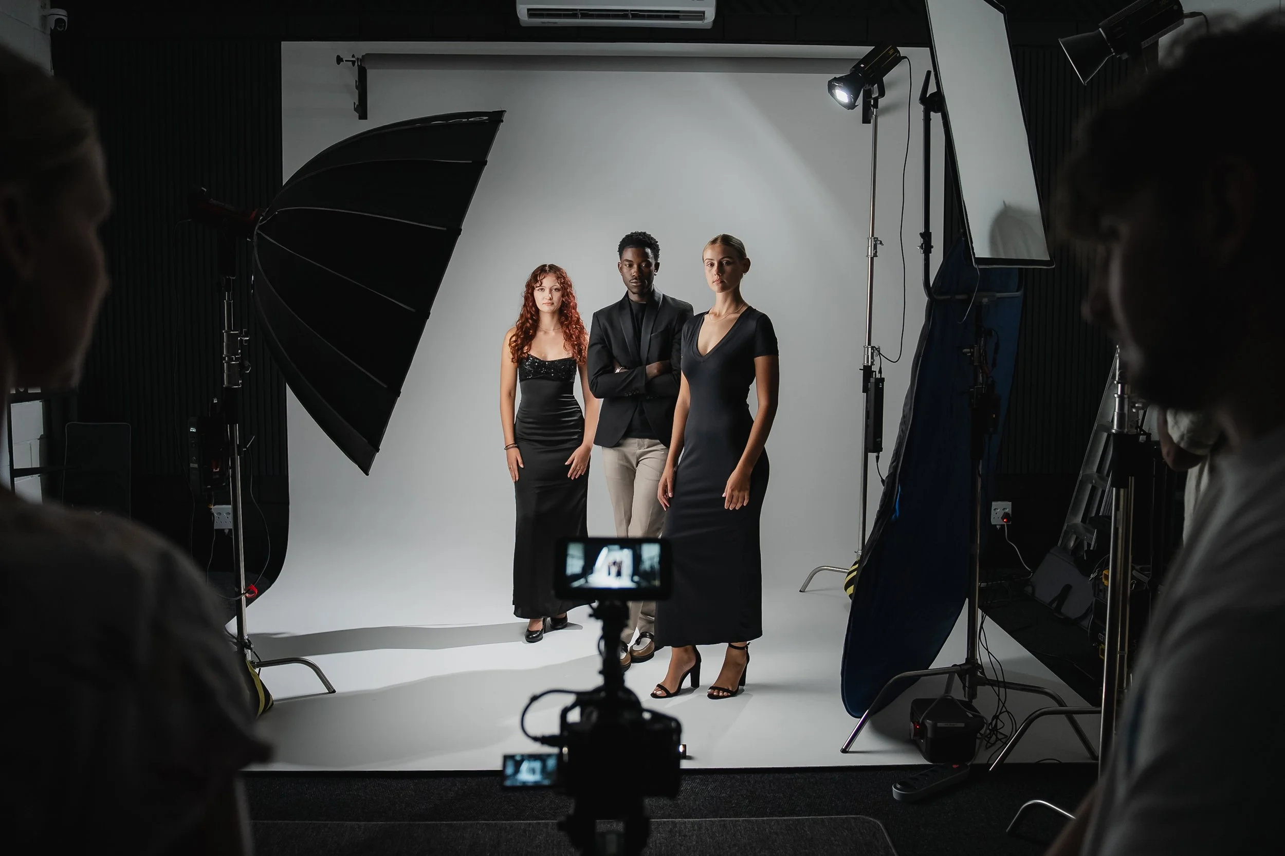 Three models standing in front of a gray backdrop during a photoshoot, with professional lighting and crew members visible around them.