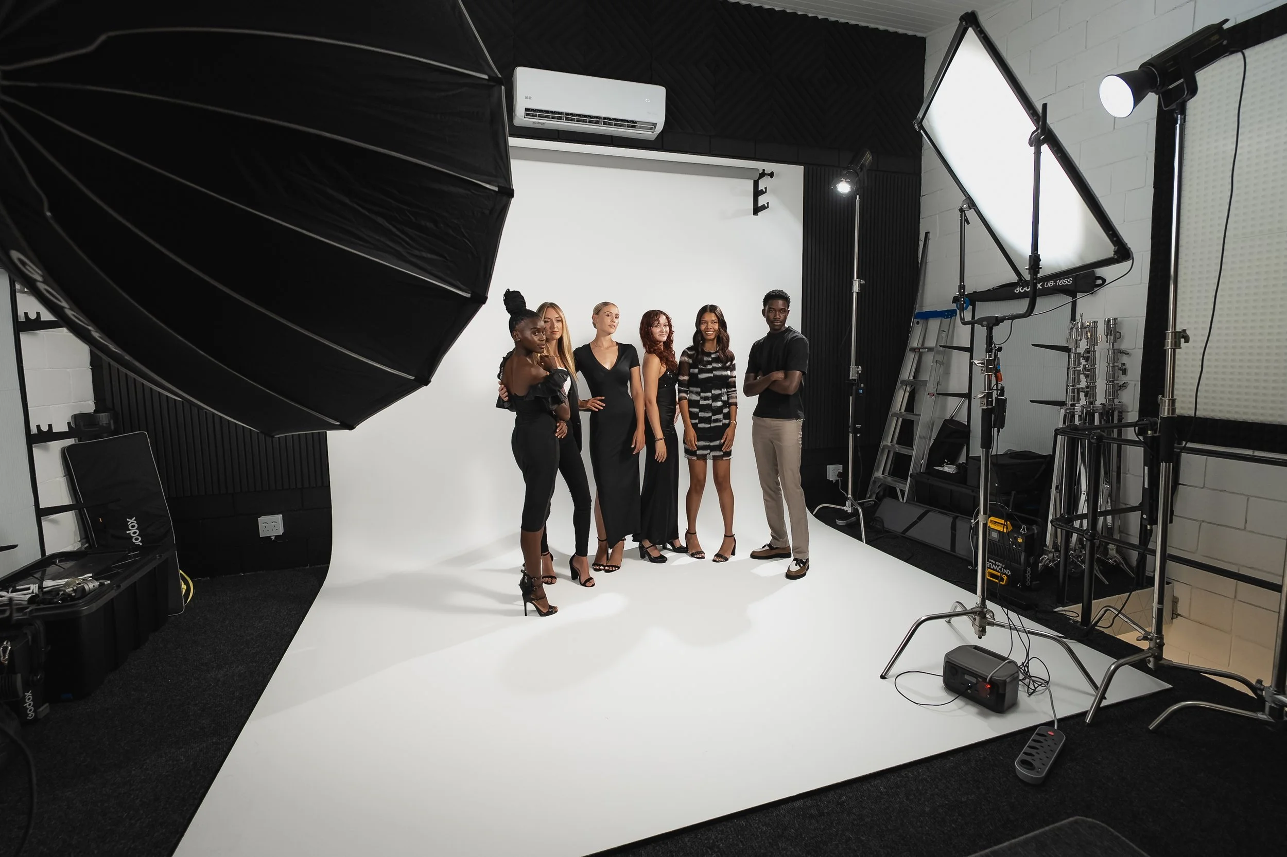 Group of five diverse models in fashionable black outfits posing in a photography studio with professional lighting equipment and a white backdrop.