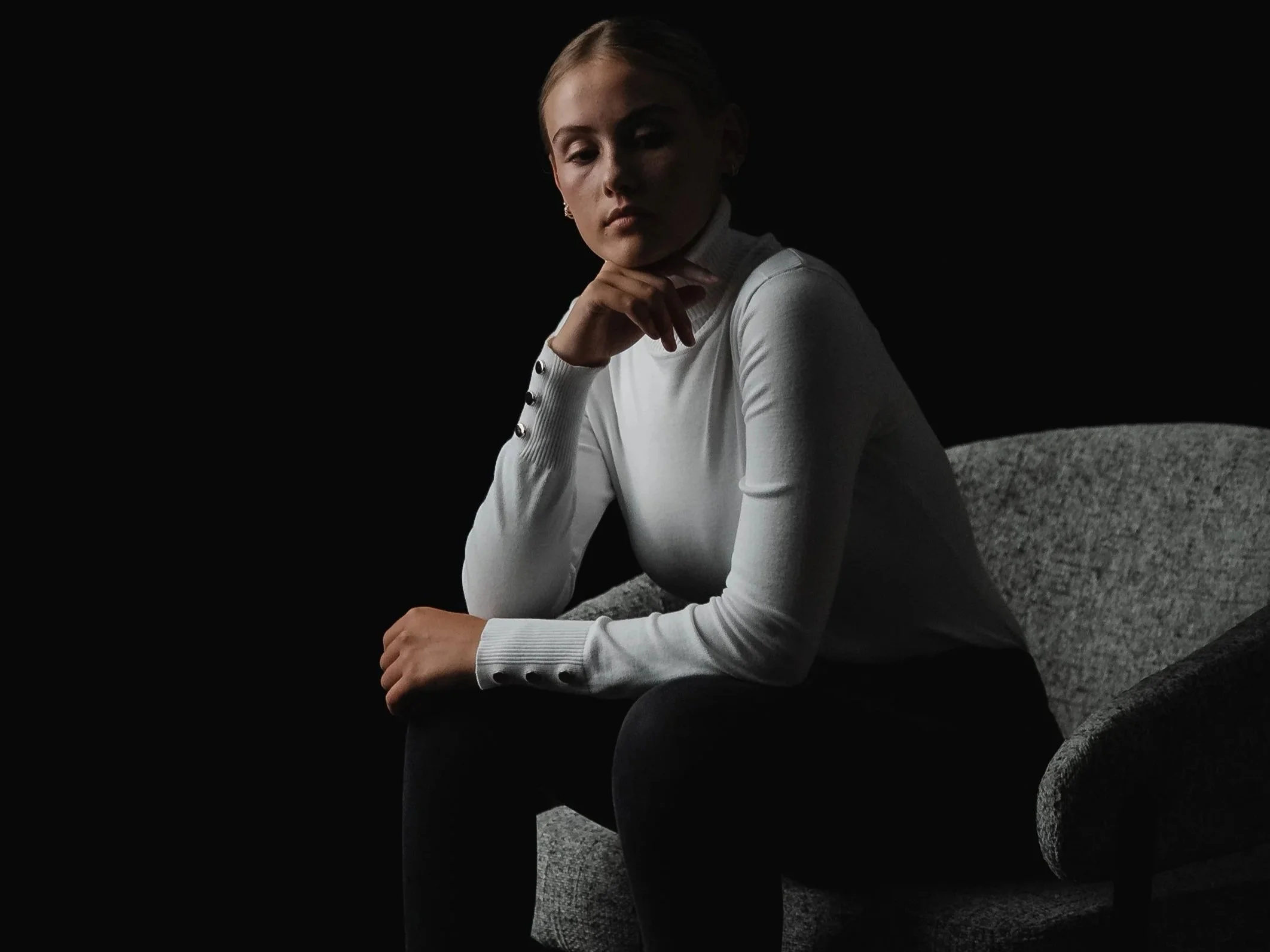A woman with blonde hair wearing a white long-sleeve top sitting on a gray armchair against a black background, with a contemplative expression.