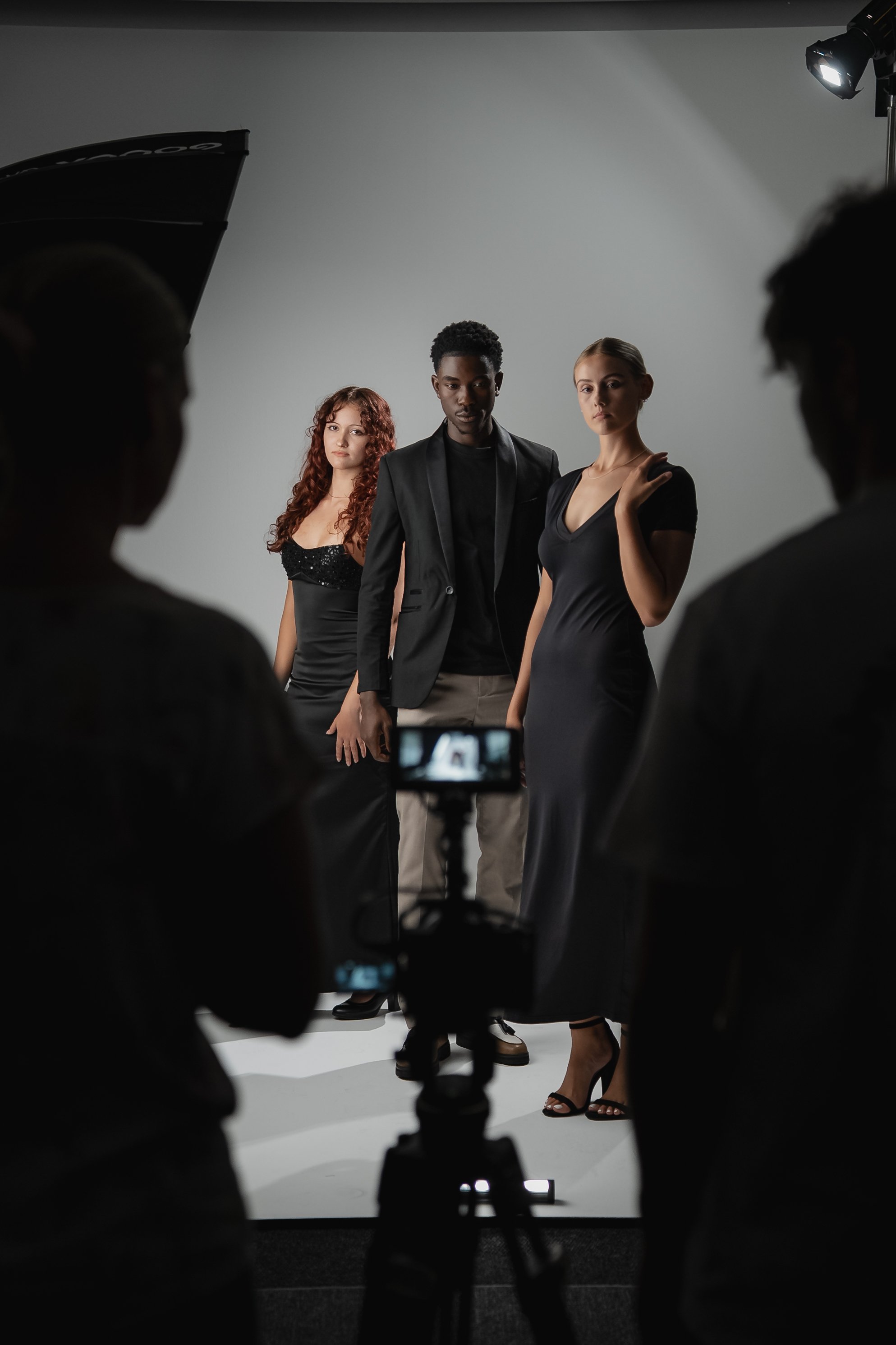 Three models standing in front of a gray backdrop during a photoshoot, with photographers and studio equipment visible.