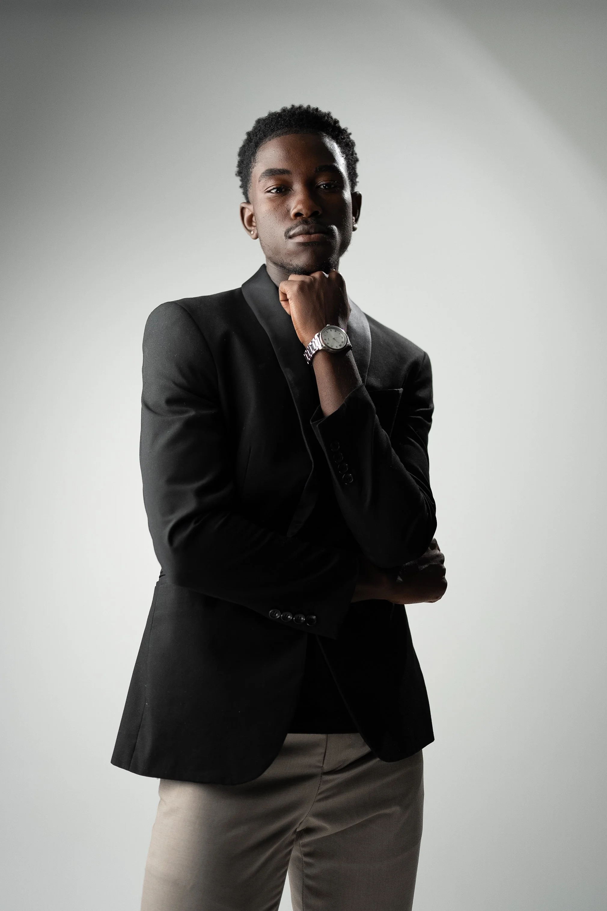 A young man wearing a black blazer, light-colored pants, and a wristwatch, posing with his hand near his face against a neutral background.