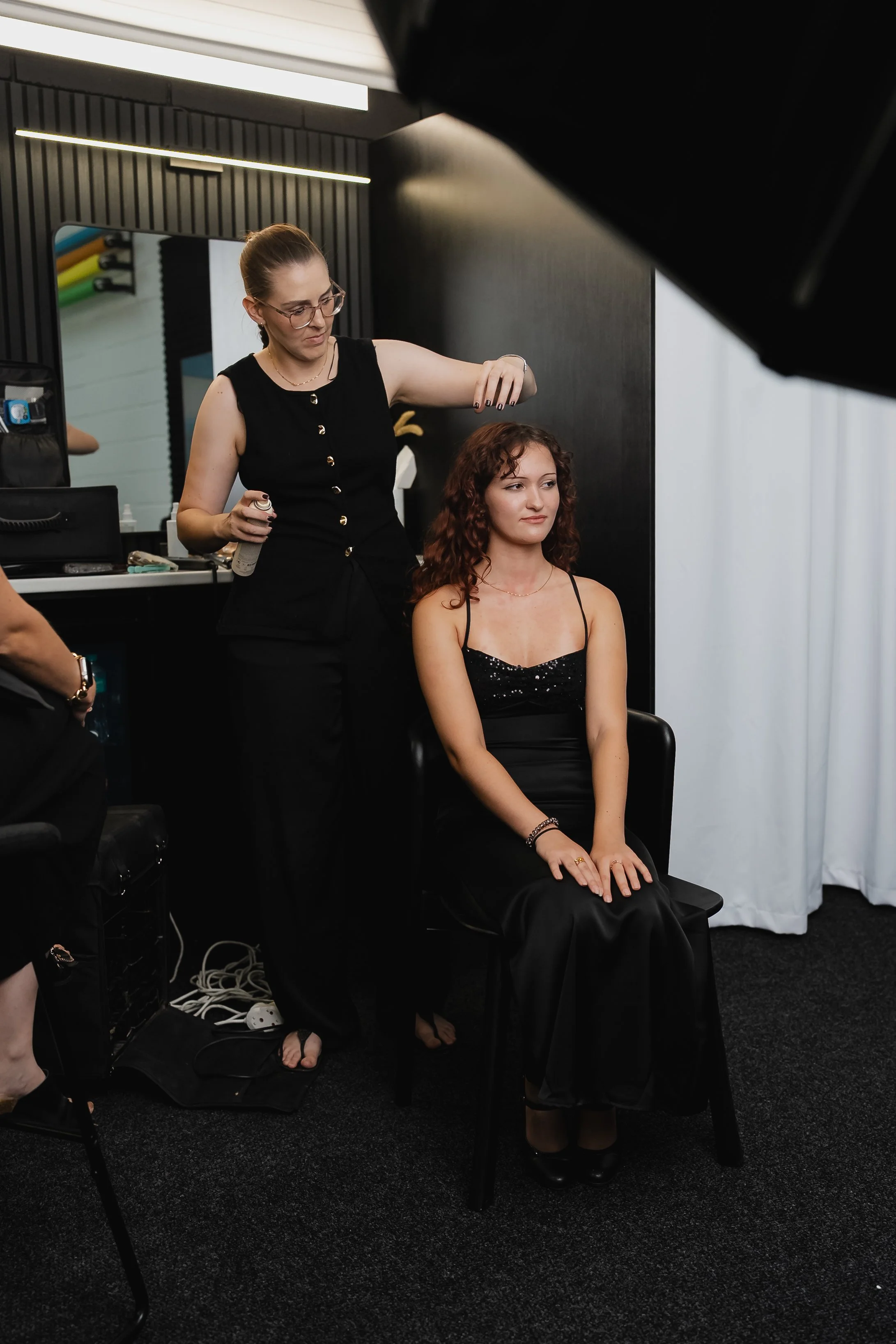 A woman getting her hair styled by a hairdresser in a salon, sitting on a chair wearing a black dress with spaghetti straps.