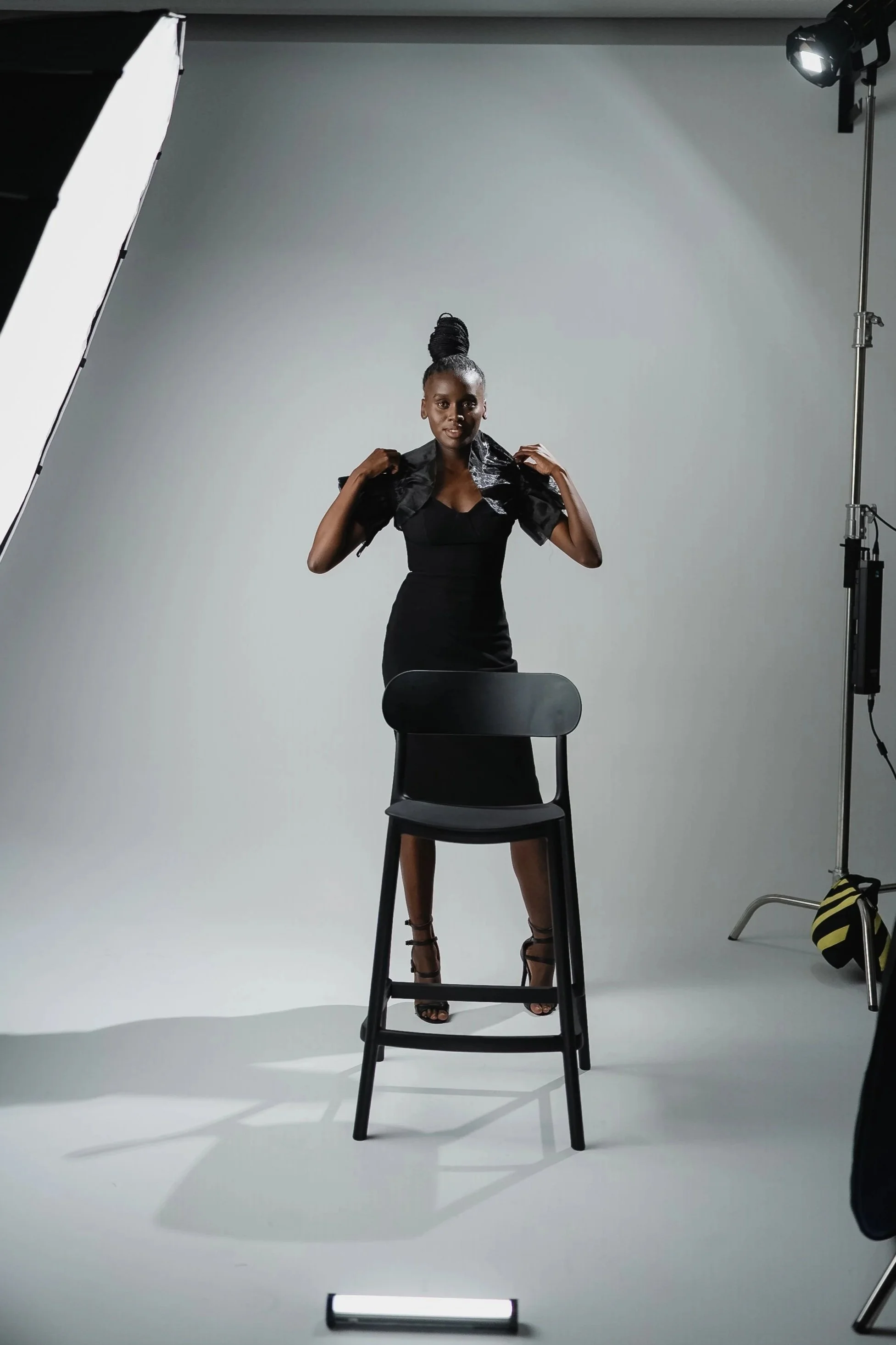 A woman standing on a black chair in a photography studio with a white backdrop, adjusting her black jacket, wearing a black dress and high heels, with professional lighting and photography equipment around her.