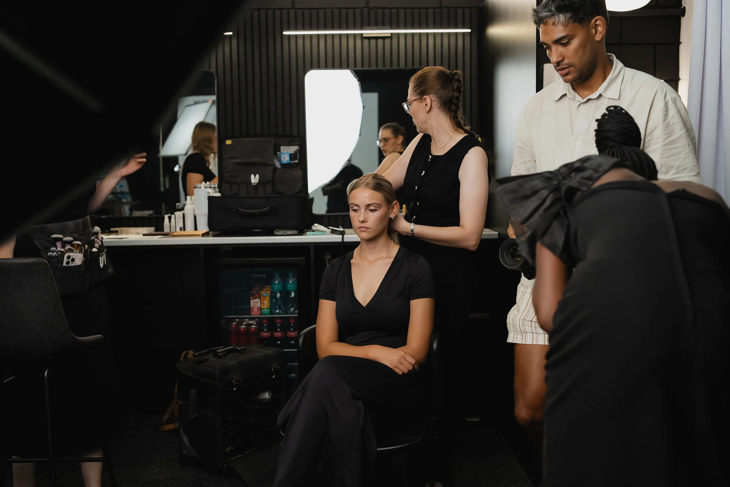 A makeup artist applies makeup to a seated woman in a black shirt, while a photographer documents the process in a professional studio with lighting equipment and makeup supplies.