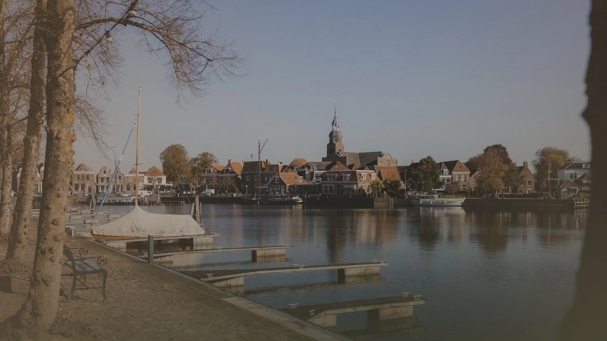 A calm river with boats docked along a shoreline, lined with trees and quaint houses in a small town with a church steeple in the background.