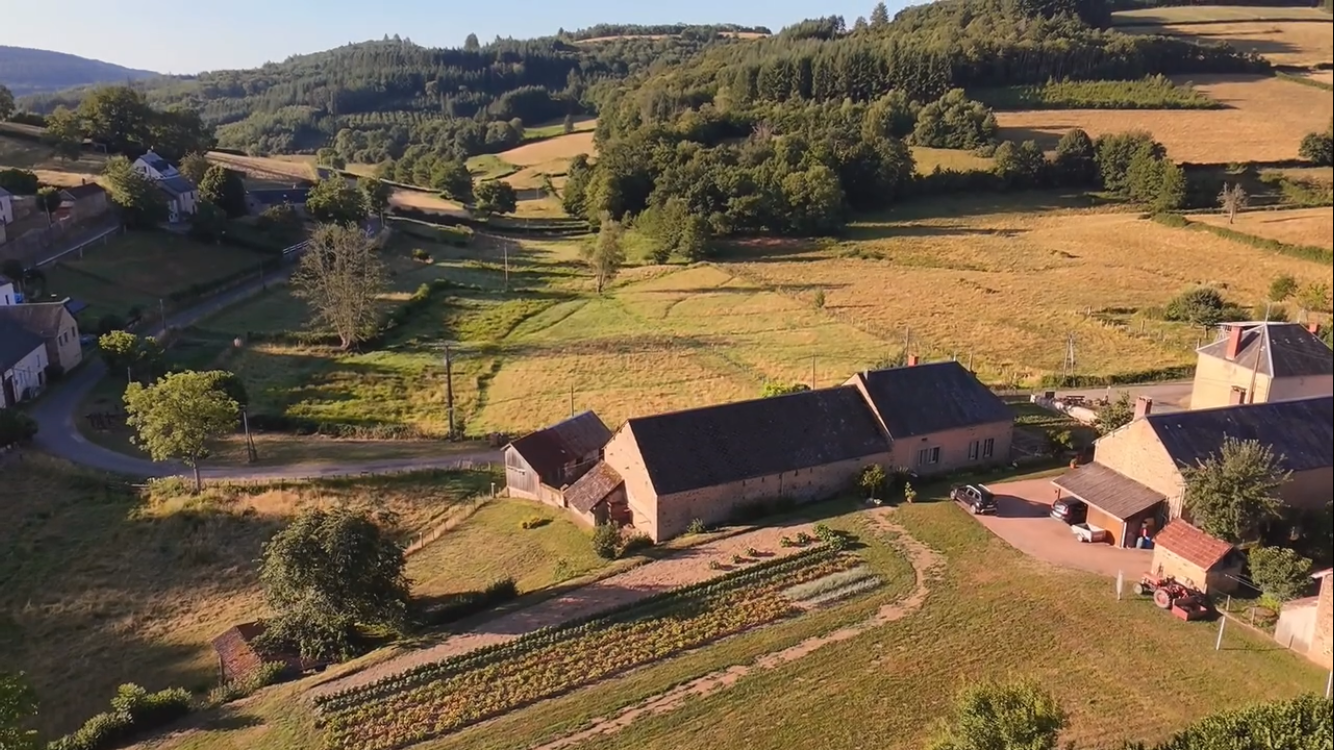 Aerial view of a rural landscape with houses, farm buildings, trees, grassy fields, and rolling hills in the background.