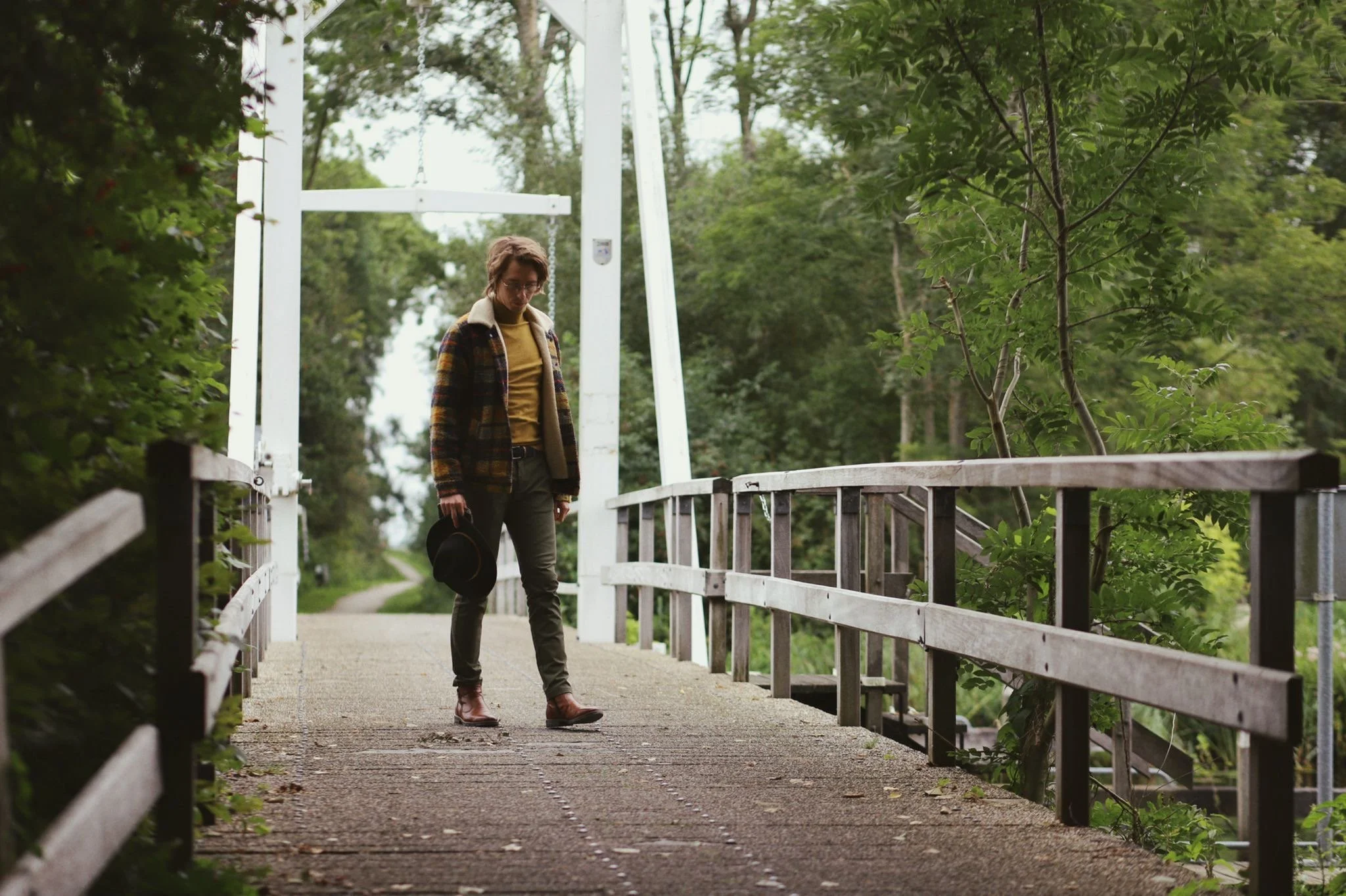 A woman walking on a small wooden bridge surrounded by green trees, holding a black hat in her right hand, dressed in a plaid jacket, yellow shirt, gray pants, and brown boots.