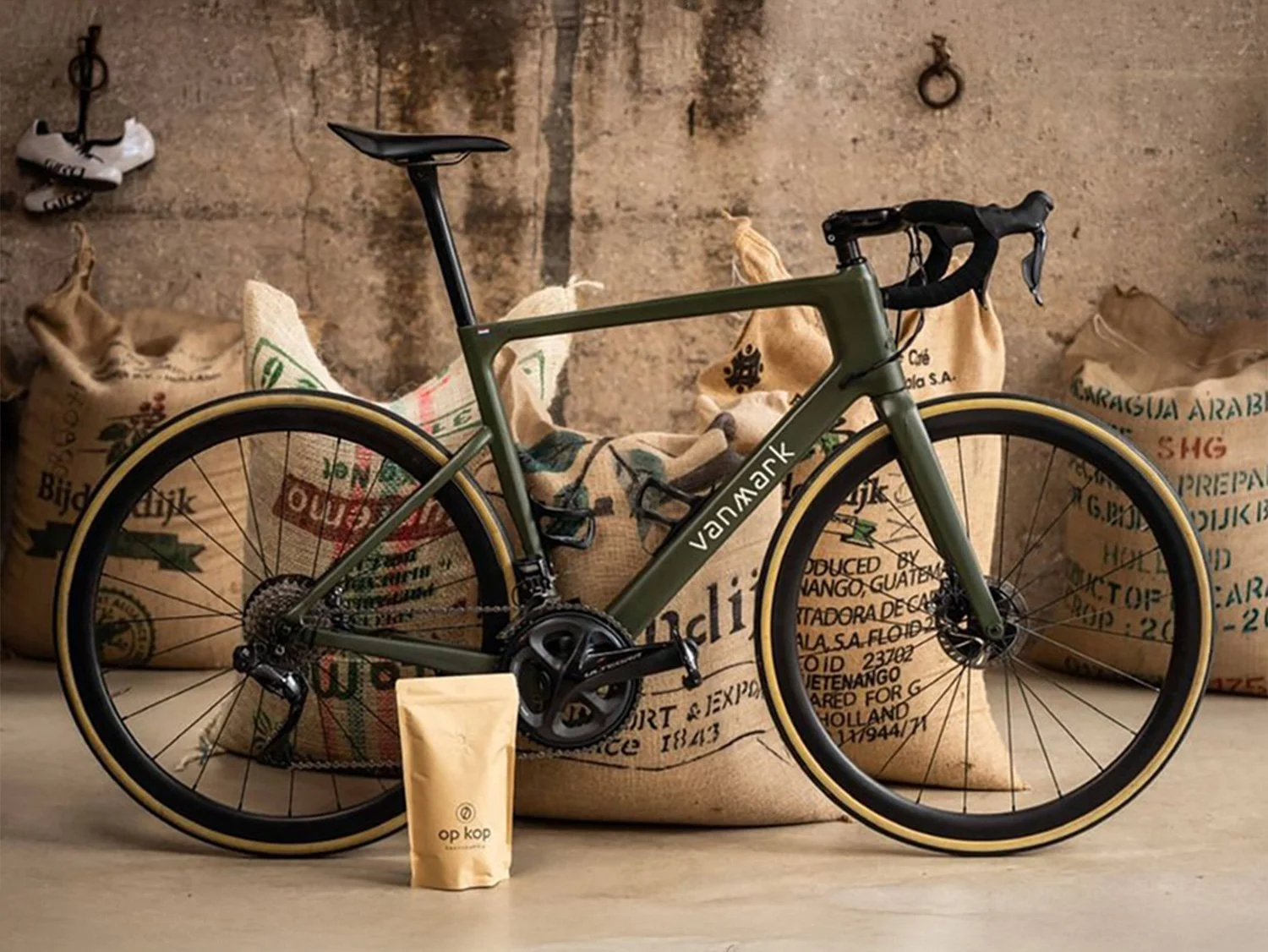 A green Vanmoof road bicycle with black handlebars and seat, positioned in front of large burlap sacks filled with coffee beans, against a rustic, textured wall. A small beige bag of coffee labeled 'OP KOP' is placed in front of the bicycle.