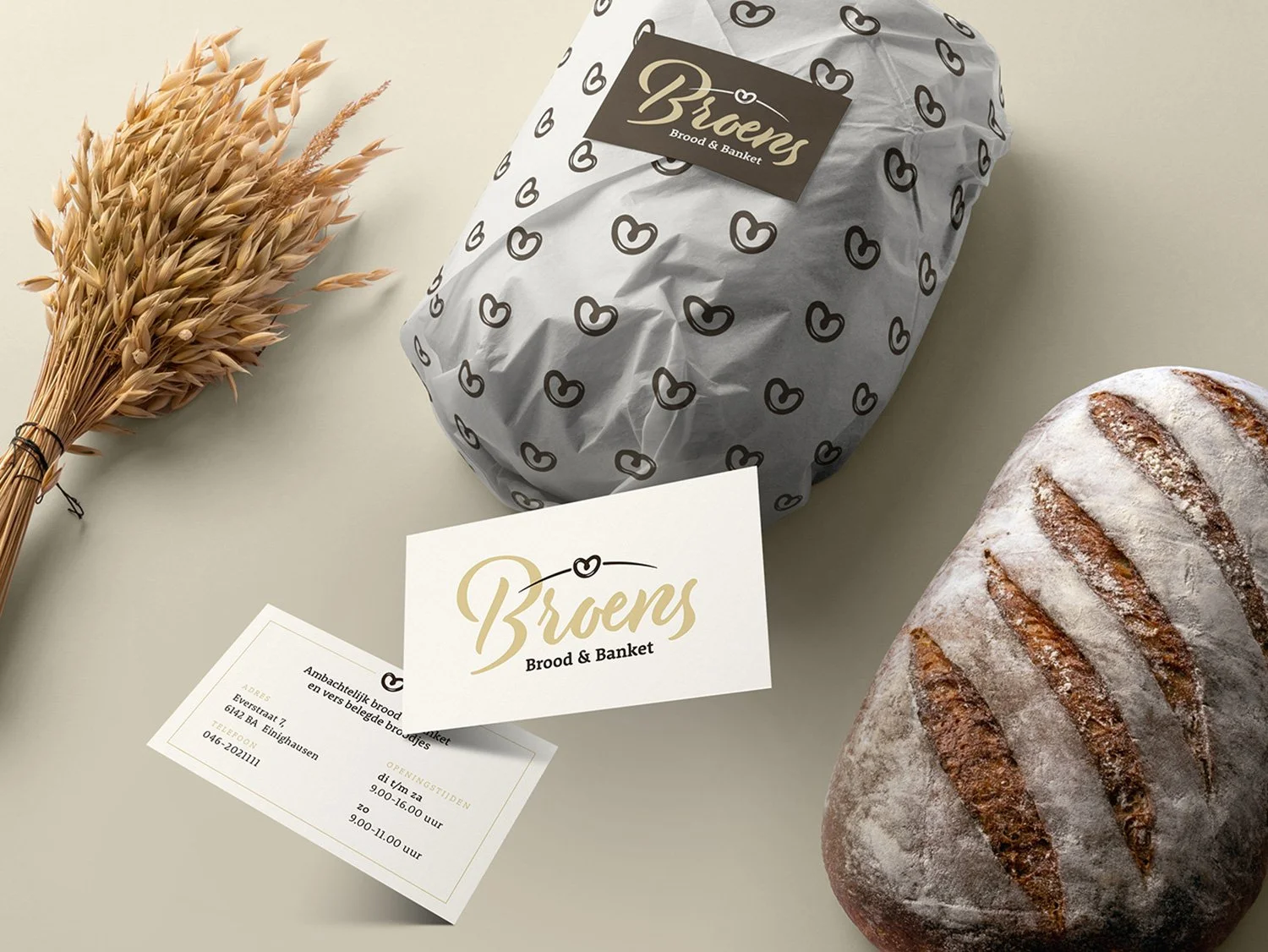 A loaf of crusty bread with grill marks, wrapped bread in paper with a black and gold logo, a business card for Briens Brood & Banket, and a bunch of dried wheat stalks