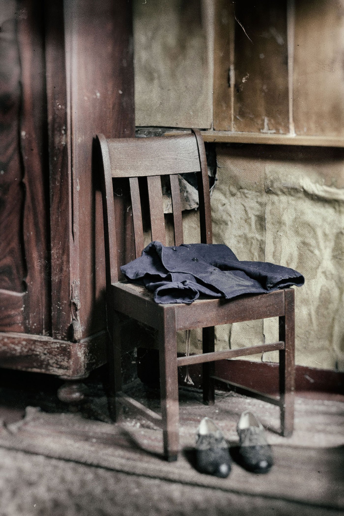 Old wooden chair with a black coat on it, pair of shoes underneath, in a dusty, worn room with wooden and stone walls.