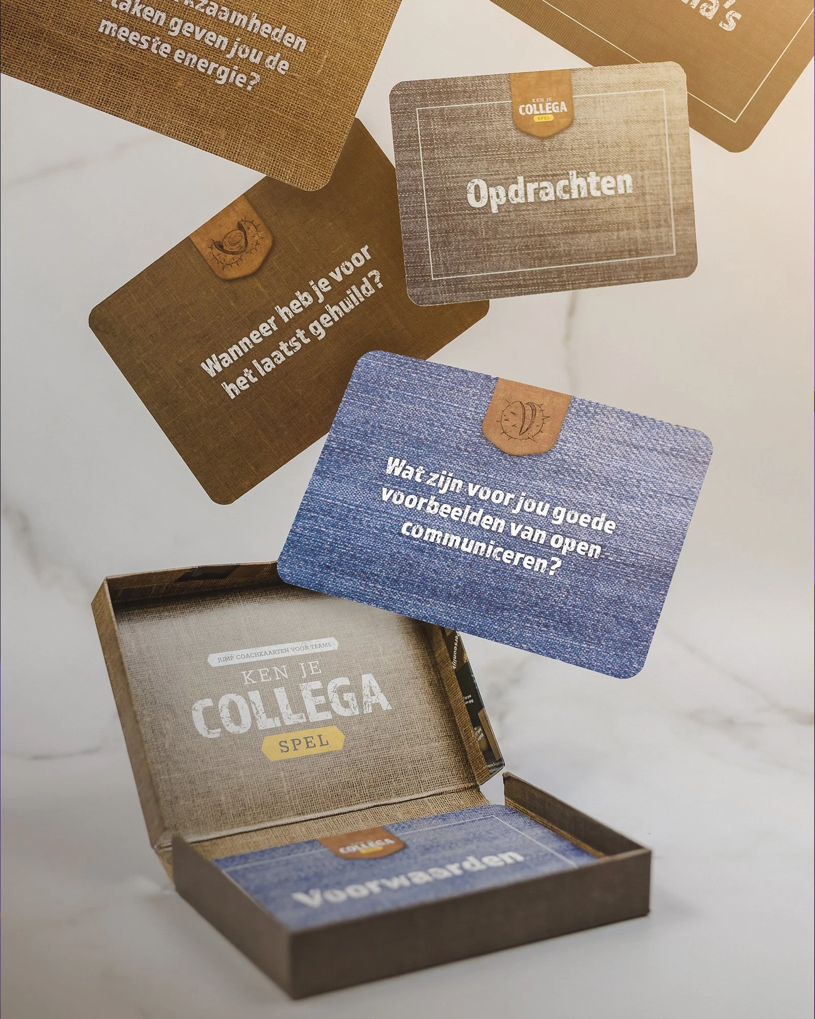 A card game box titled 'Ken je Collega?' with several cards, including one asking 'Wanneer heb je voor het laatst geluisterd?' and another asking 'Wat zijn voor jou goede voorbeelden van open communiceren?' placed above and beside the box. The box an