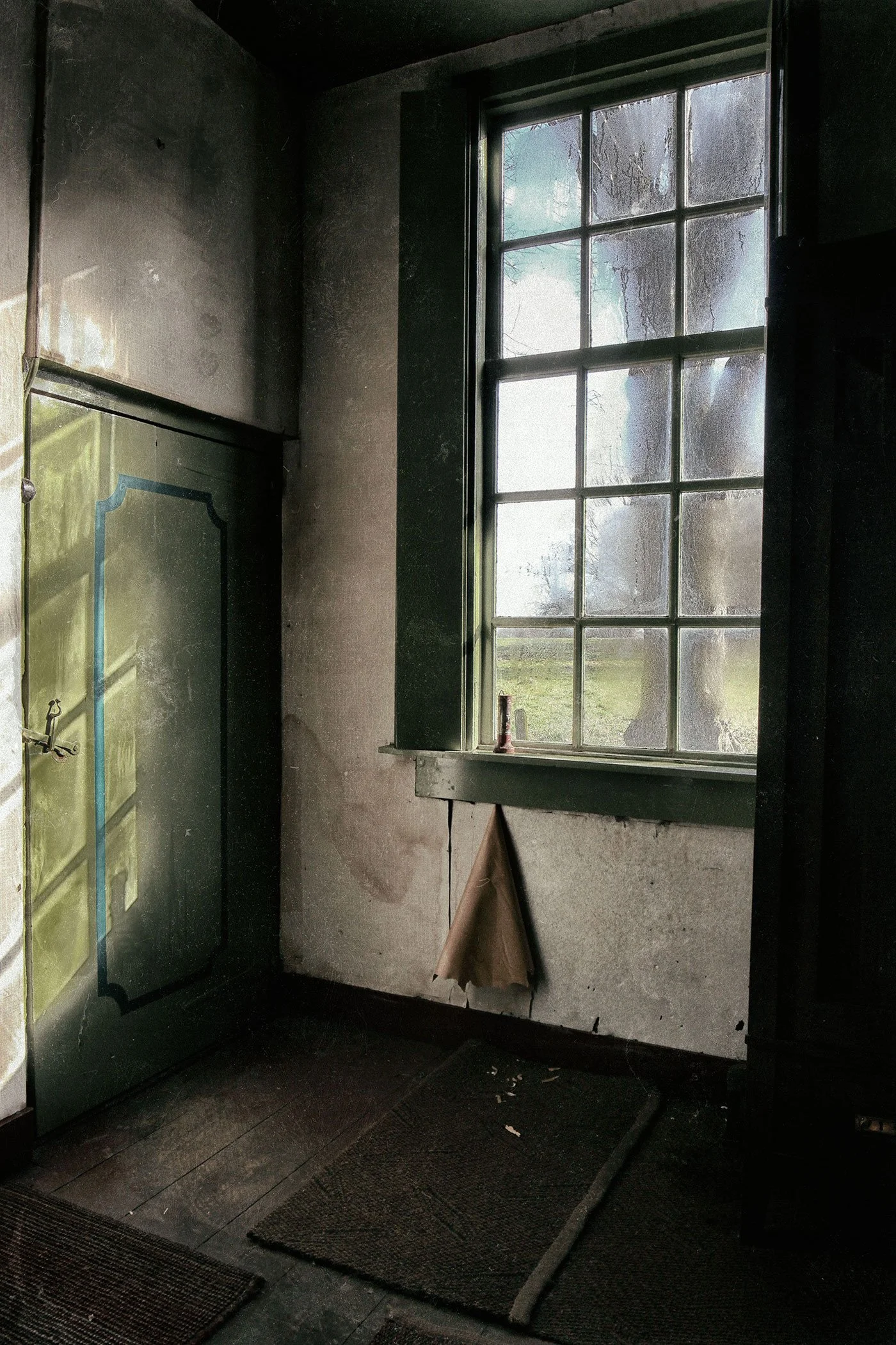 Dimly lit room with a large window showing an outdoor landscape with trees and grass. Inside, there's a green door with a clock and a key hook, a brown paper bag hanging on the wall, and a dark floor with some mats.