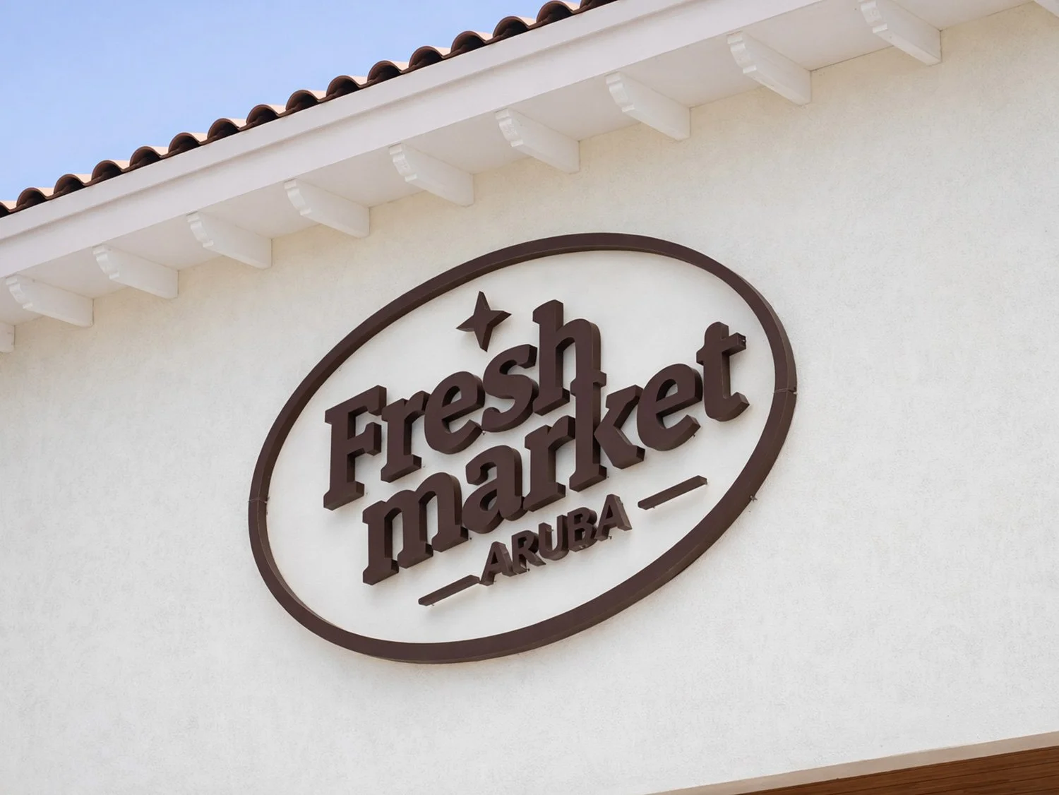 Fresht Market Aruba sign on the exterior wall of a building, featuring a white background and a brown oval outline, with brown 3D text and a star.