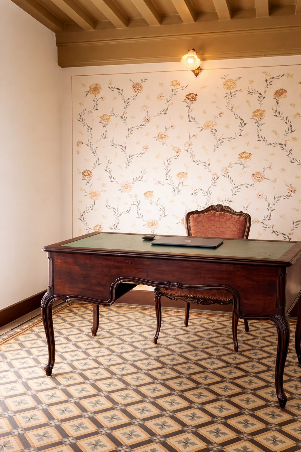 A vintage wooden desk with curved legs and a laptop and mouse on top, against a floral wallpapered wall with a classic chair behind the desk, in a room with patterned tile flooring and a wall-mounted light fixture.