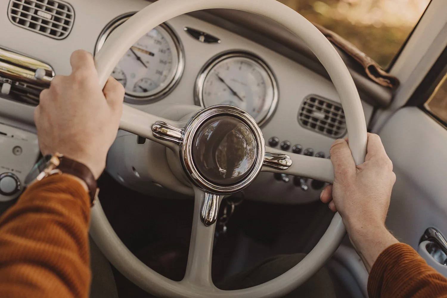 A person wearing an orange-brown long sleeve shirt is holding a vintage white steering wheel of a car, with dashboard gauges visible, including speedometer and fuel gauge.