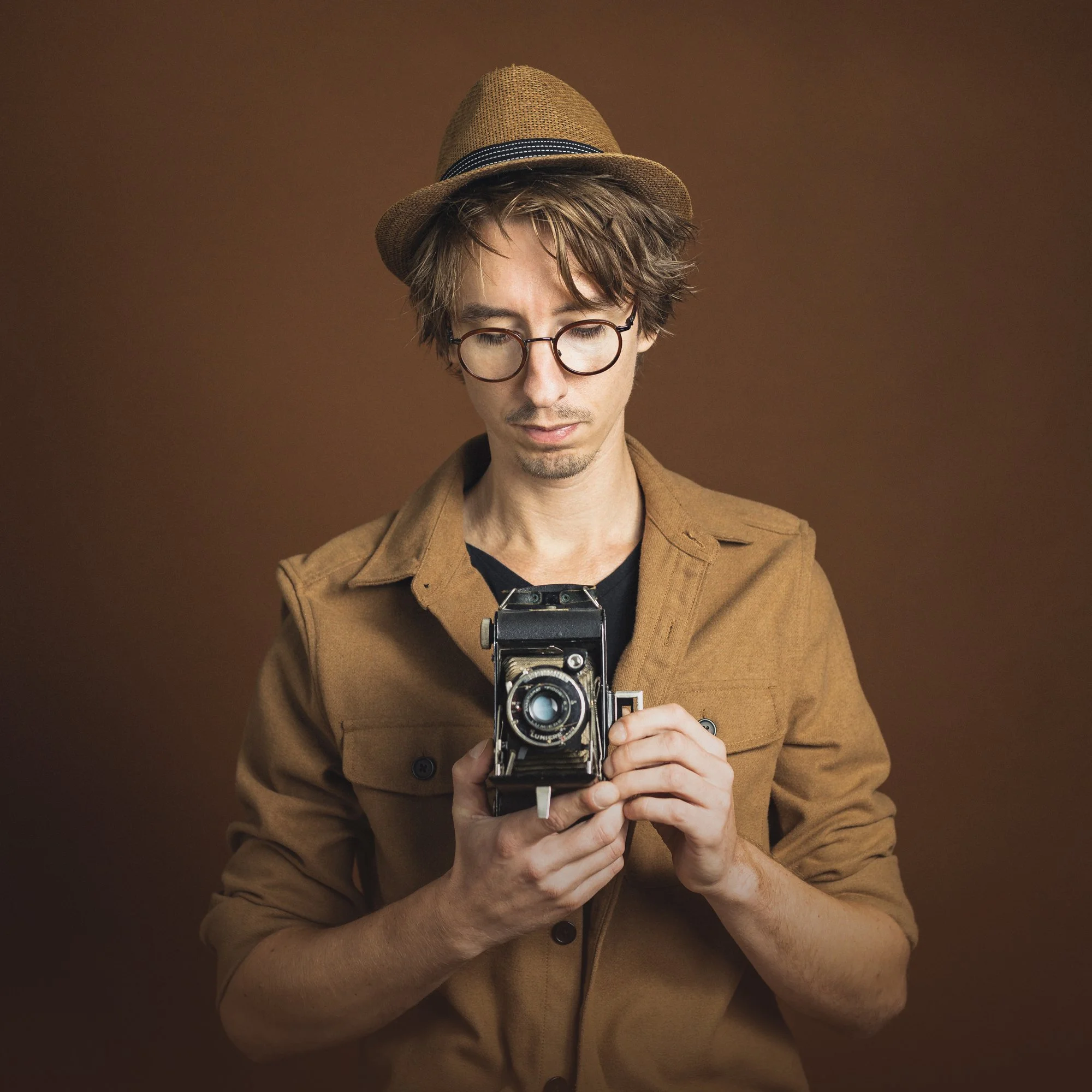 A young man with glasses and a brown jacket holding a vintage camera against a brown background.