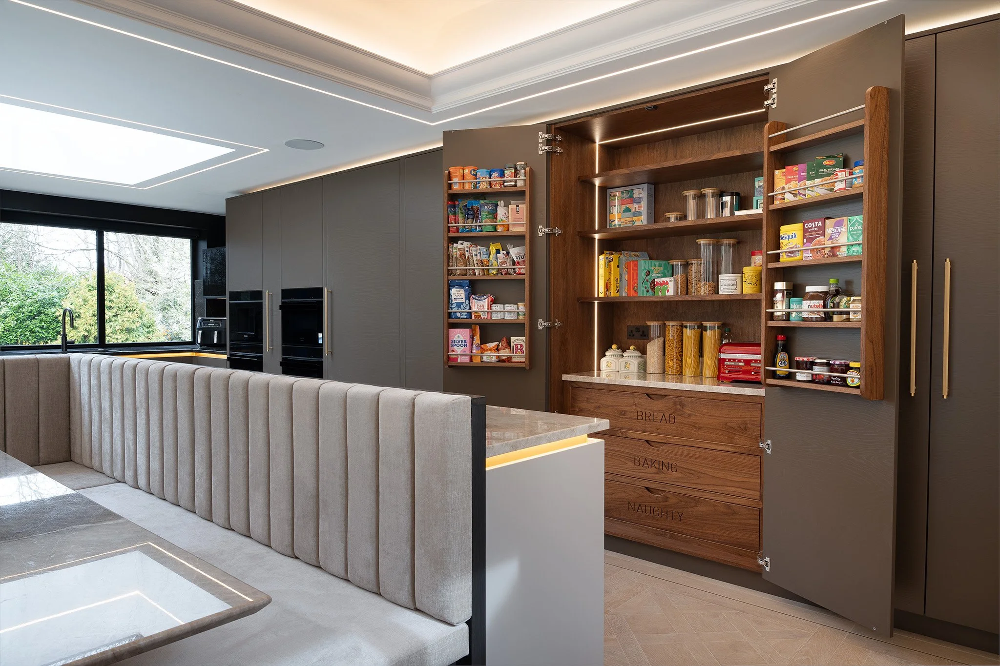 Modern kitchen with dark grey cabinets, open wooden shelves labeled 'Bread,' 'Baking,' and 'Naughtiness,' containing various food items and jars. A large window with greenery outside, beige seating, and a marble countertop.