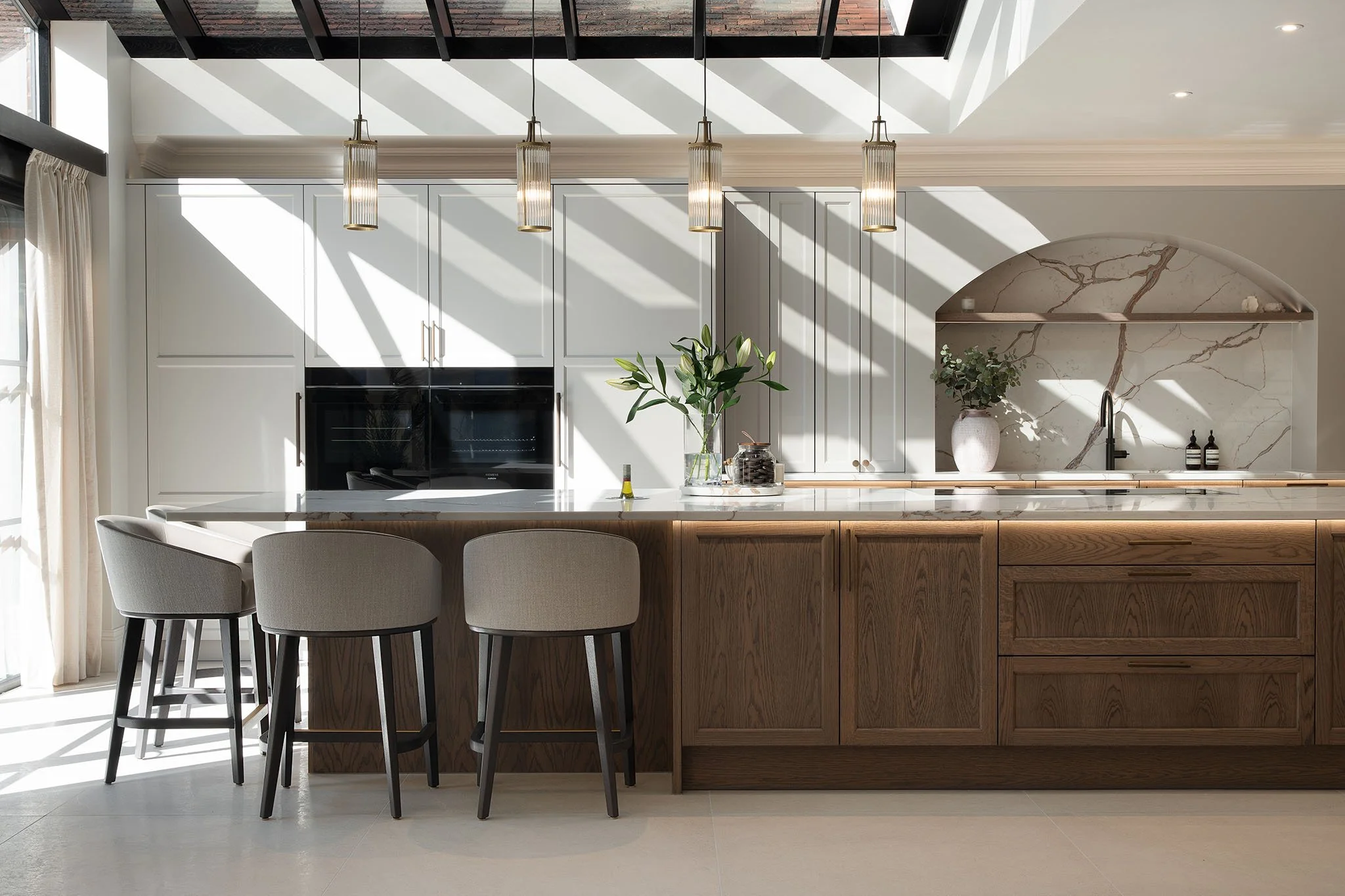 Modern kitchen with white cabinets, a marble backsplash, wood lower cabinets, a kitchen island with bar stools, pendant lights, and sunlight streaming through a skylight.