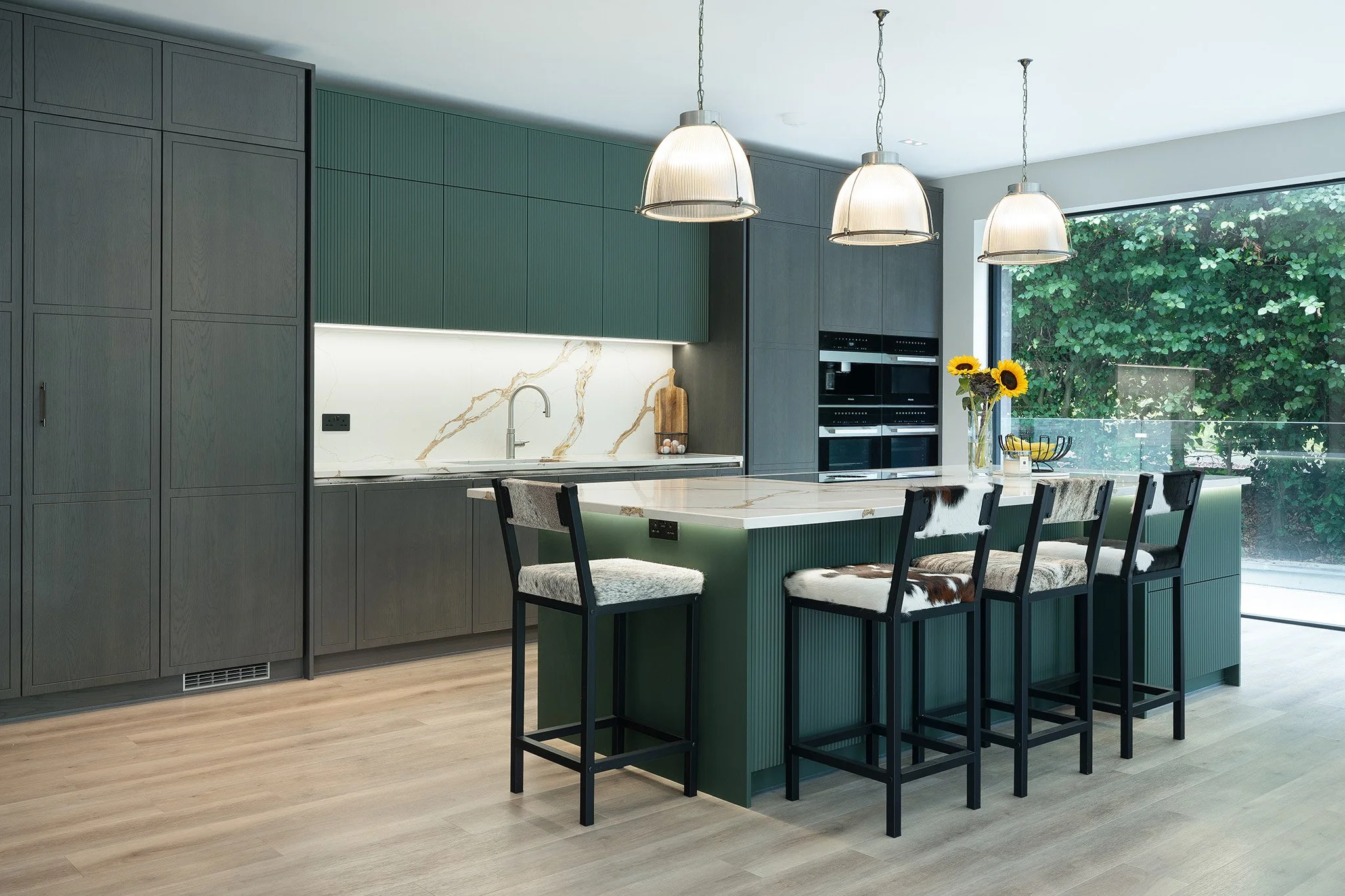 Modern kitchen with green cabinets, marble island, black bar stools with cowhide seats, hanging pendant lights, large window with greenery outside, and sunflower decor.