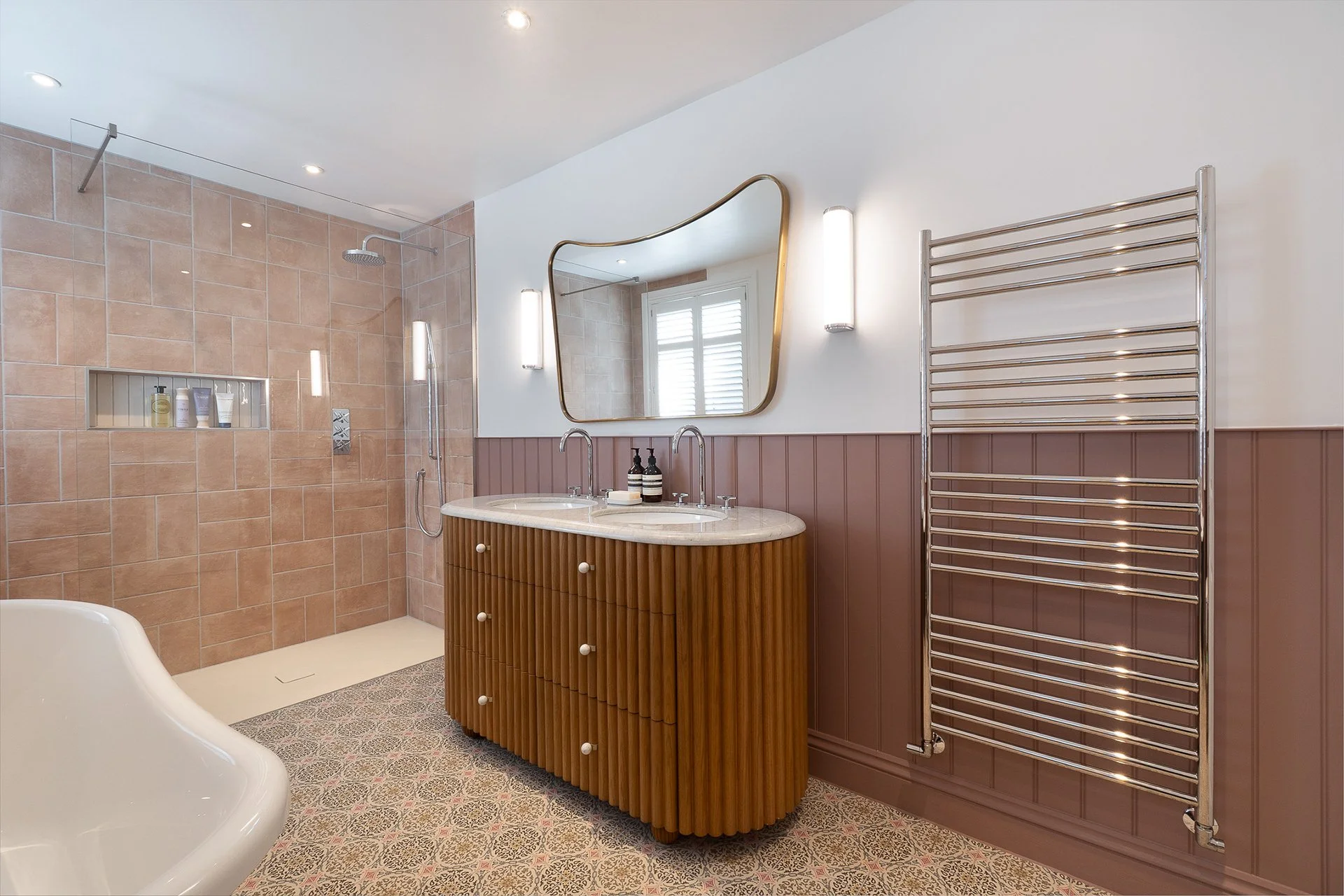 Modern bathroom featuring a walk-in shower with beige tiles, a wooden vanity with a marble countertop, a large irregularly shaped mirror, and a metallic heated towel rack. The room has stylish lighting and a patterned tile floor.