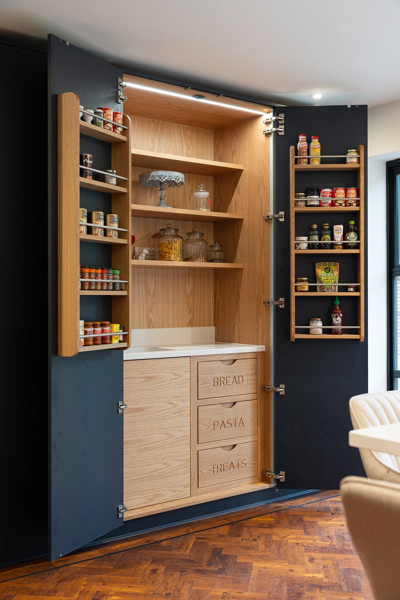 Open kitchen cabinet with wooden shelves, labeled drawers for bread, pasta, and treats, and spice racks inside doors.
