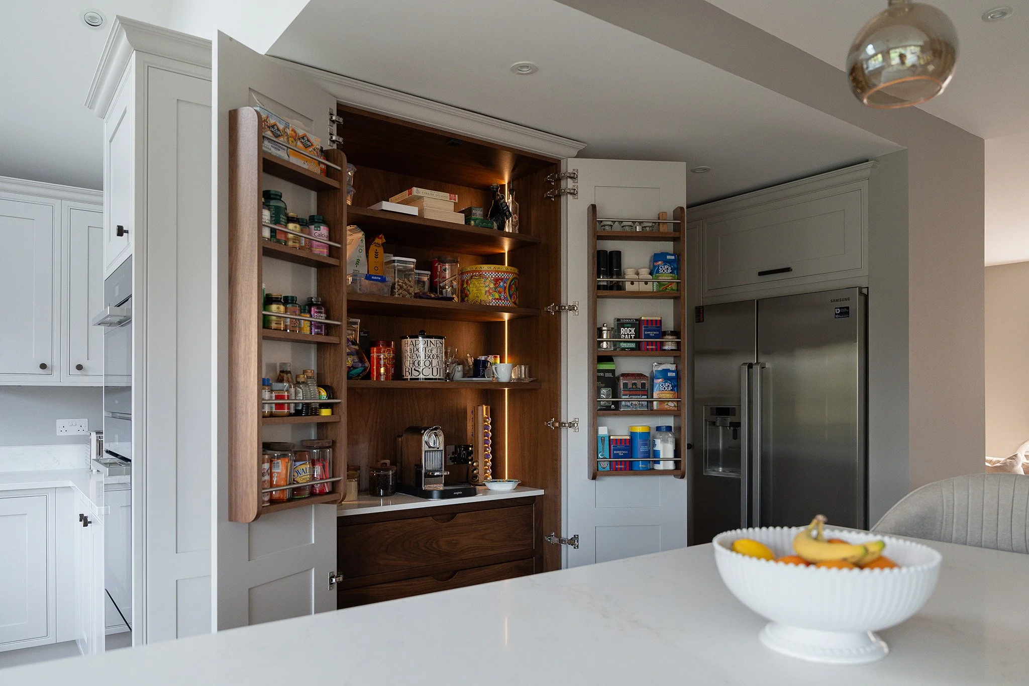 Open kitchen cabinet with food items, a coffee machine, and a bowl of fruit in the foreground.