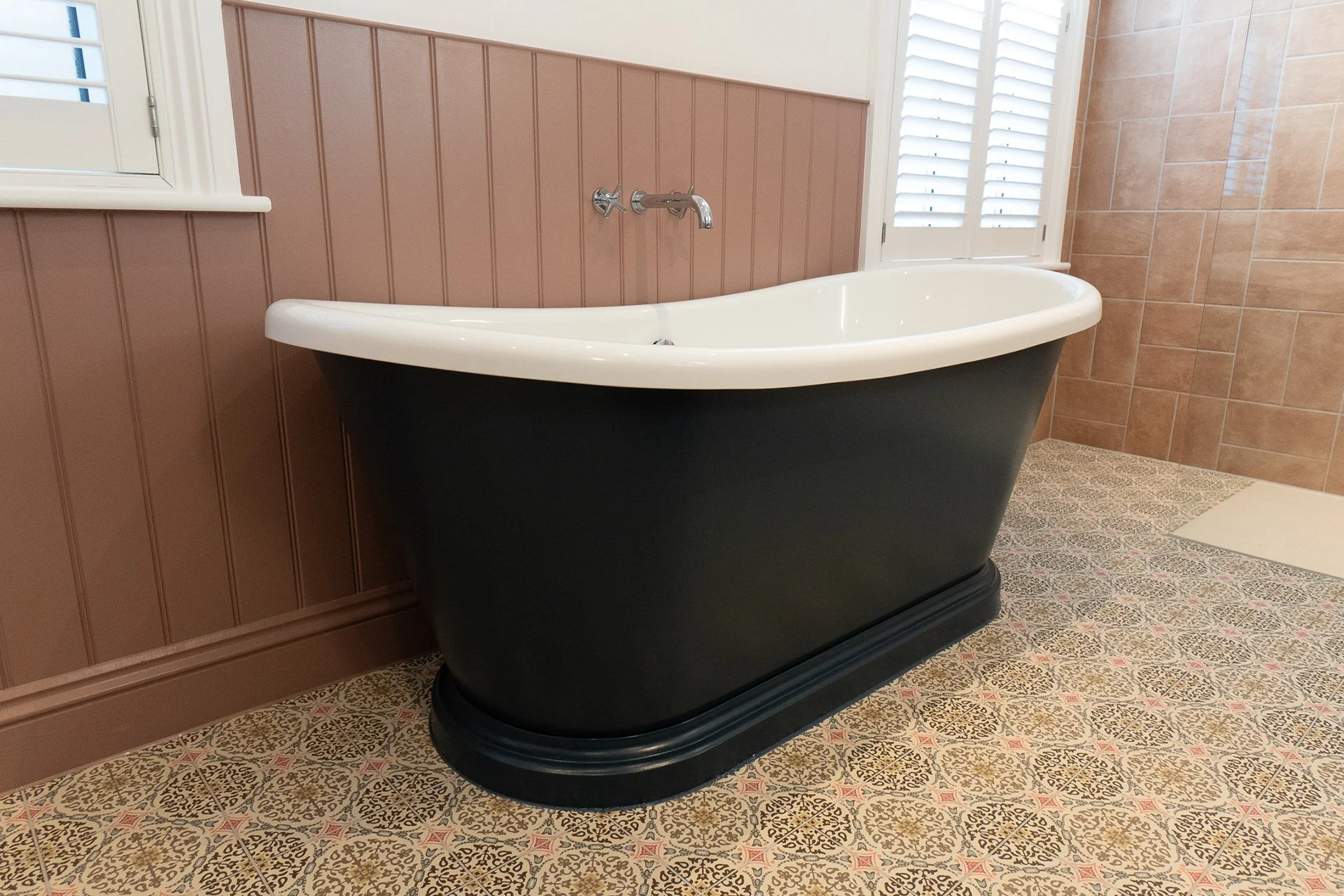 A vintage clawfoot bathtub with a black exterior and white interior, placed on a patterned tile floor in a bathroom with pink and beige wall paneling and a large window with shutters.