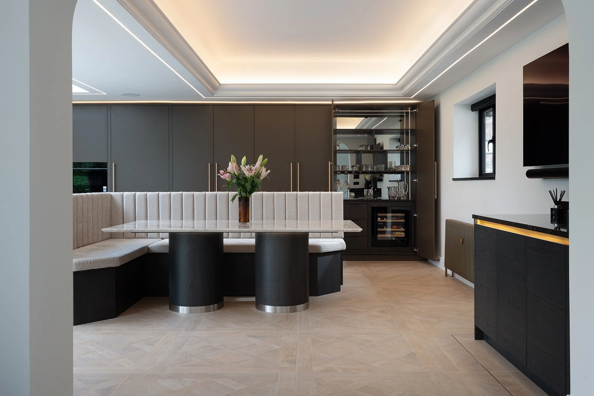 Modern dining area with a marble table, beige upholstered banquette, and a vase of pink lilies. Background features dark cabinetry, glassware cupboard, small window, and flat-screen TV on the wall.