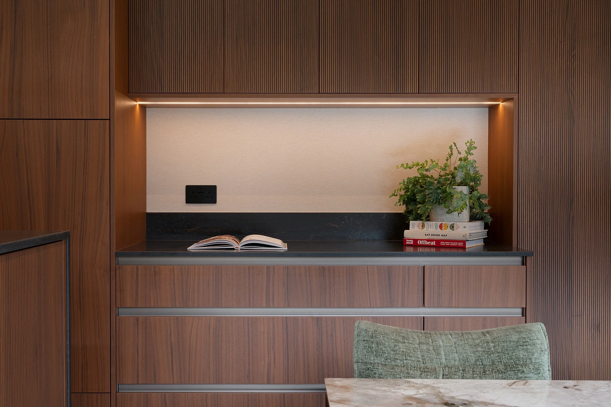 Wooden cabinet with a black countertop in a modern interior, featuring an open book, a potted plant, and a few stacked books, with a marble table and green chair in the foreground.