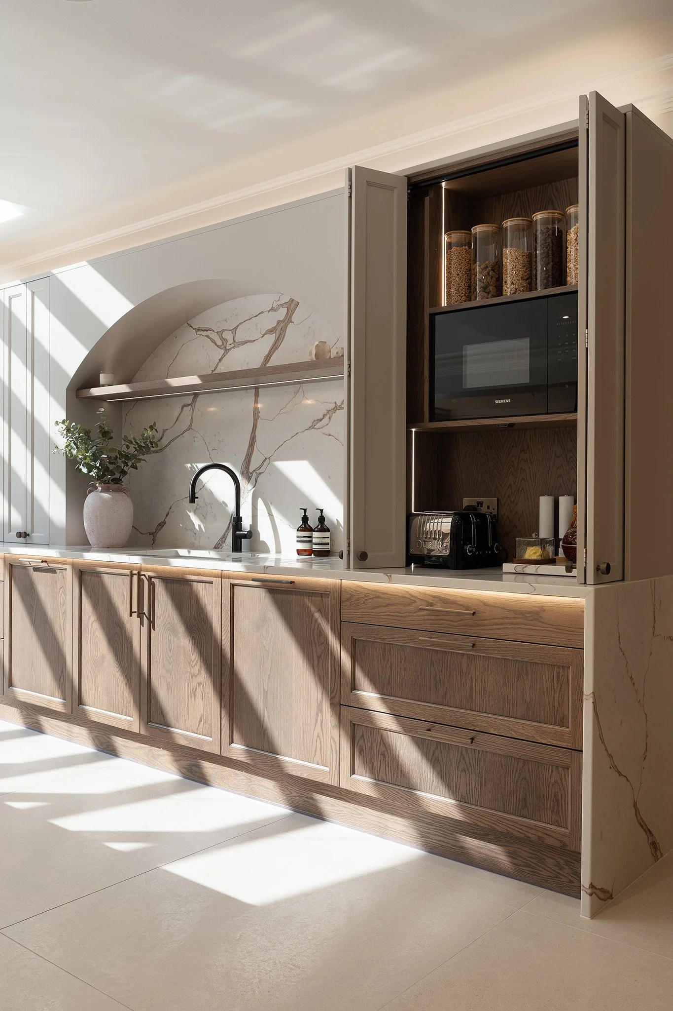 Modern kitchen with wooden cabinets, marble backsplash, open shelving, and sunlight streaming through windows.