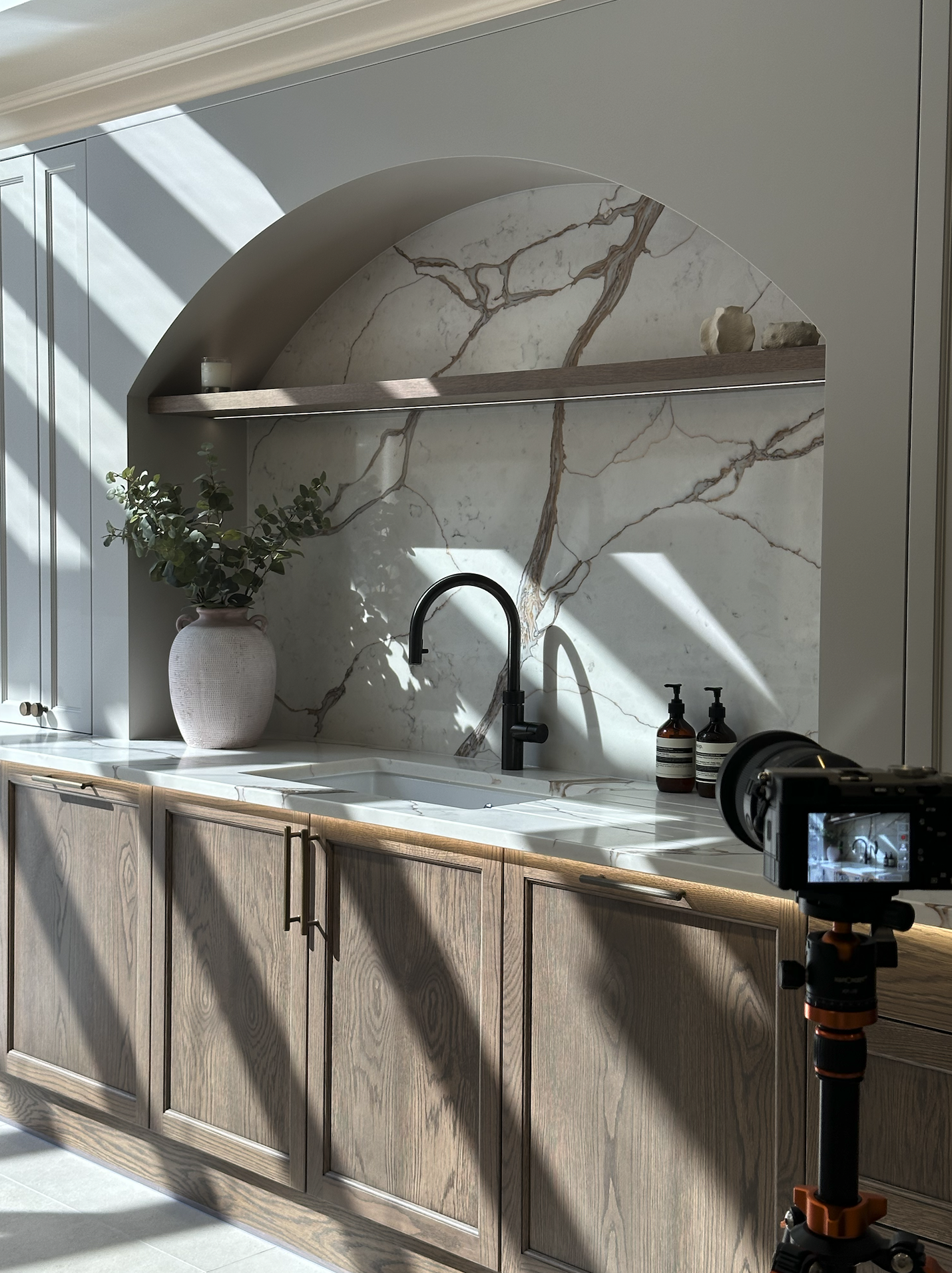 Kitchen with marble backsplash, wooden cabinets, black faucet, potted plant in a pink vase, bottles of soap, and a camera on a tripod.
